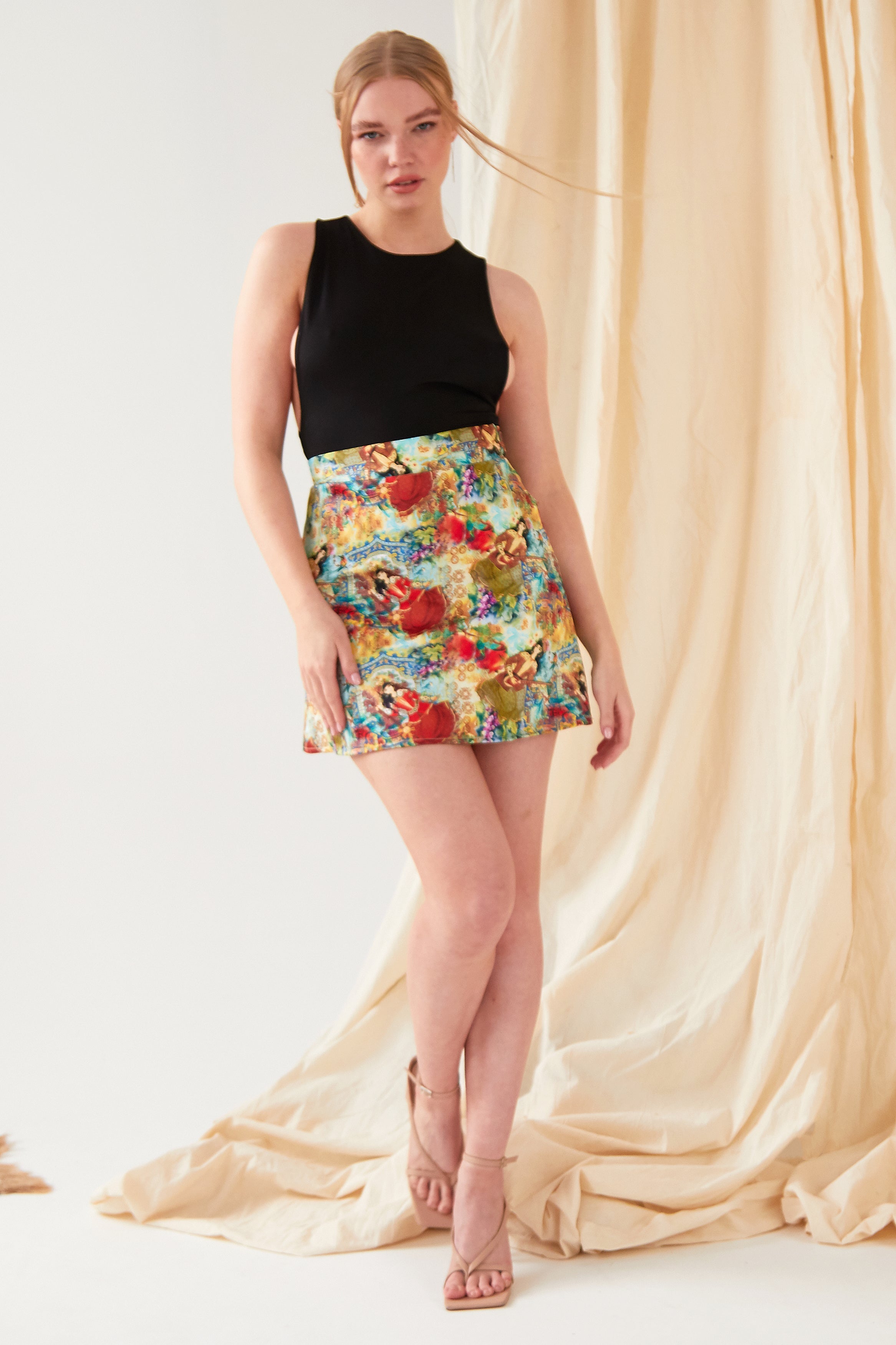Woman wearing a colorful floral skirt and black top in front of beige curtains.