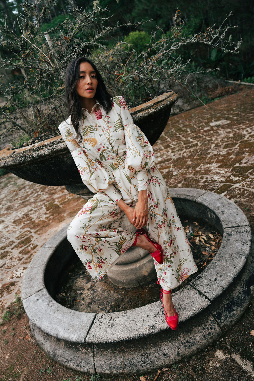 Woman in a floral silk outfit sitting in a stone fountain with a natural background.