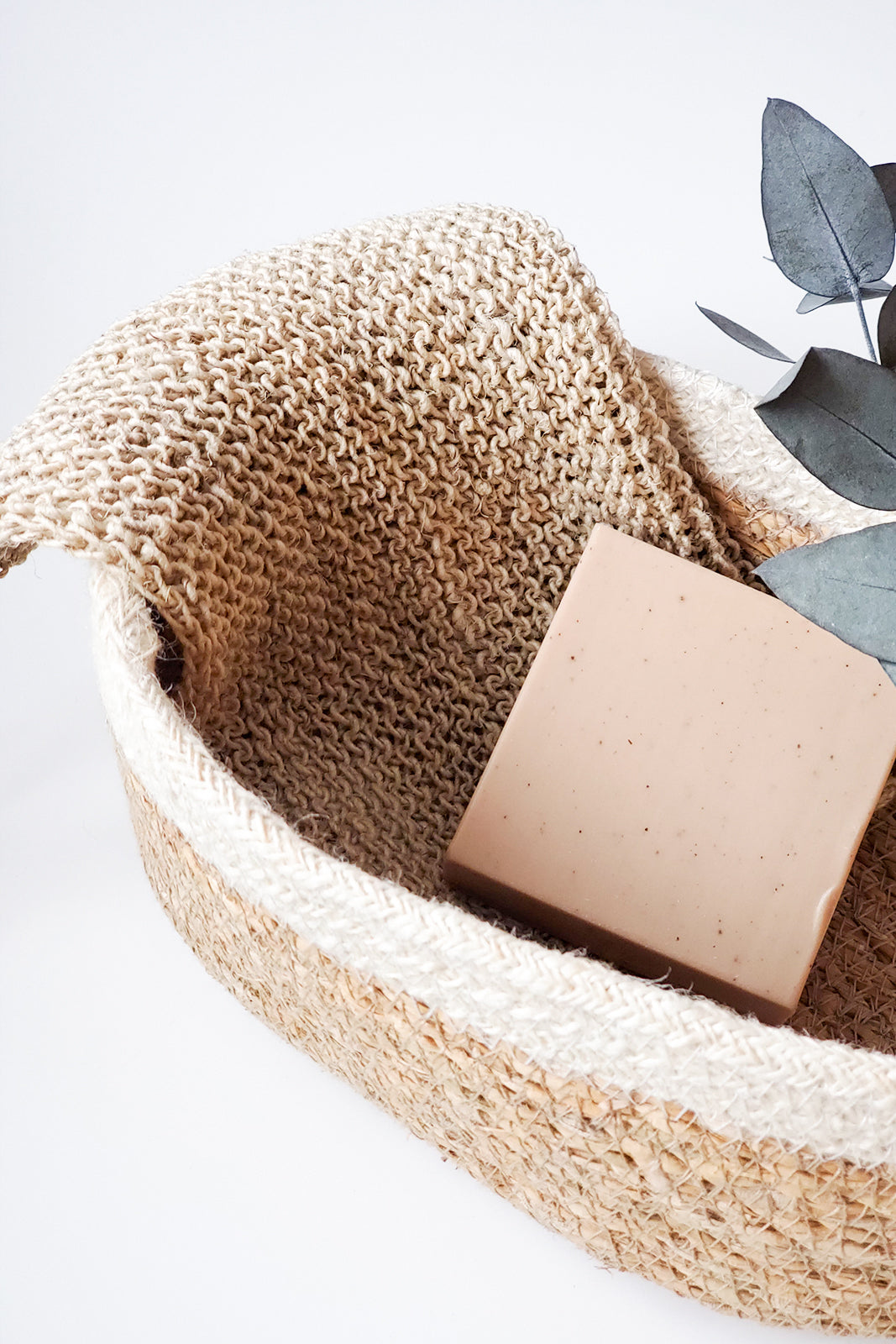 Bar of soap in a woven basket with a textured cover and decorative leaves on a light background