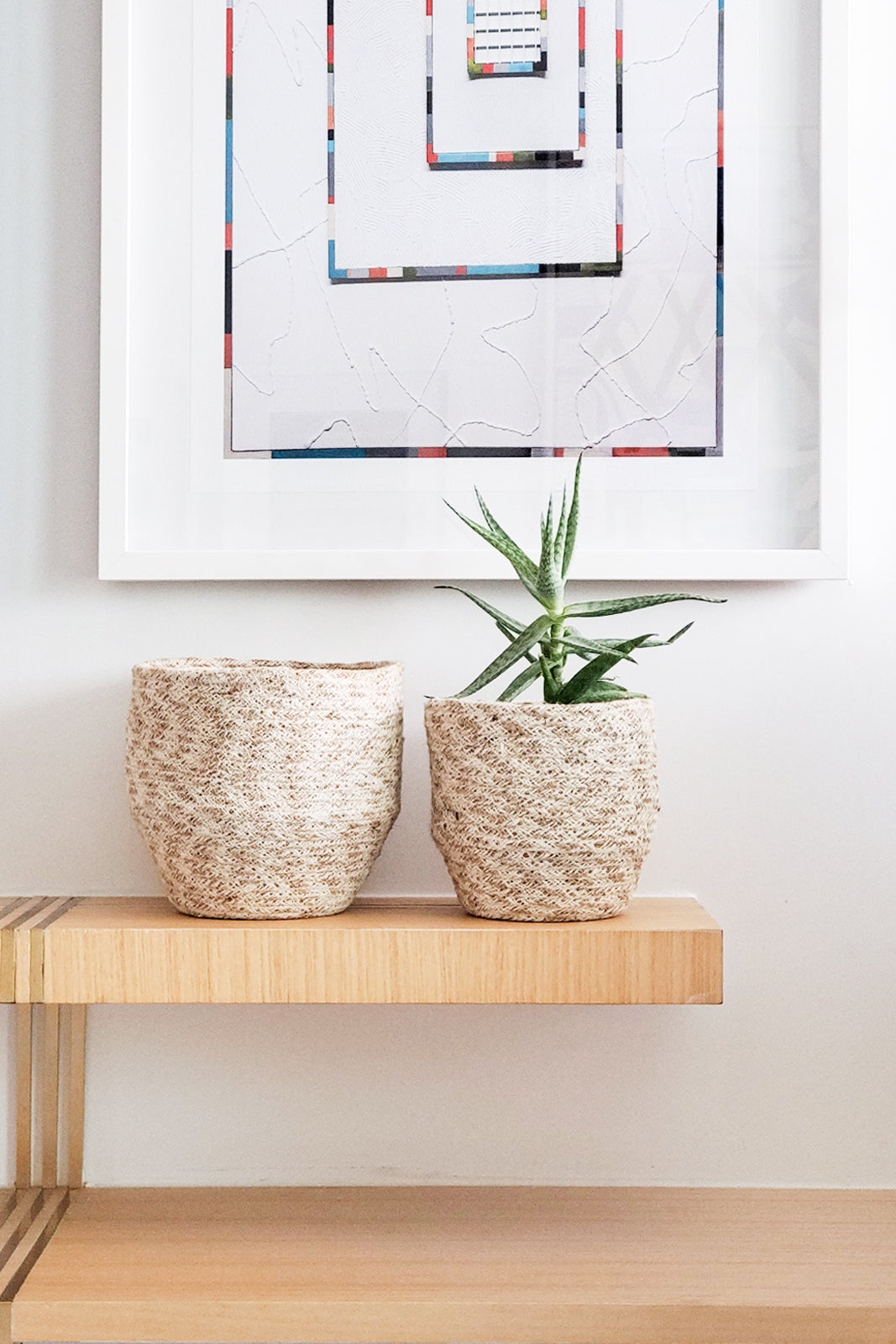 Two woven planters on a wooden shelf with a framed abstract painting above.