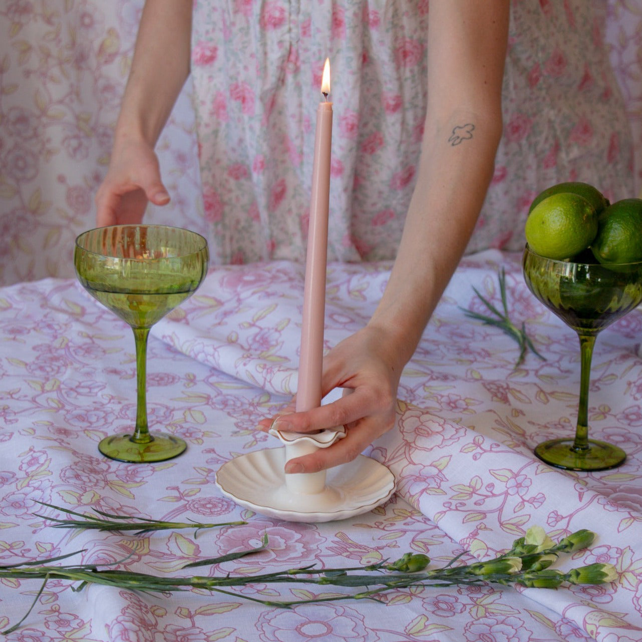 Person lighting a pink candle on a table with green glassware and floral tablecloth.