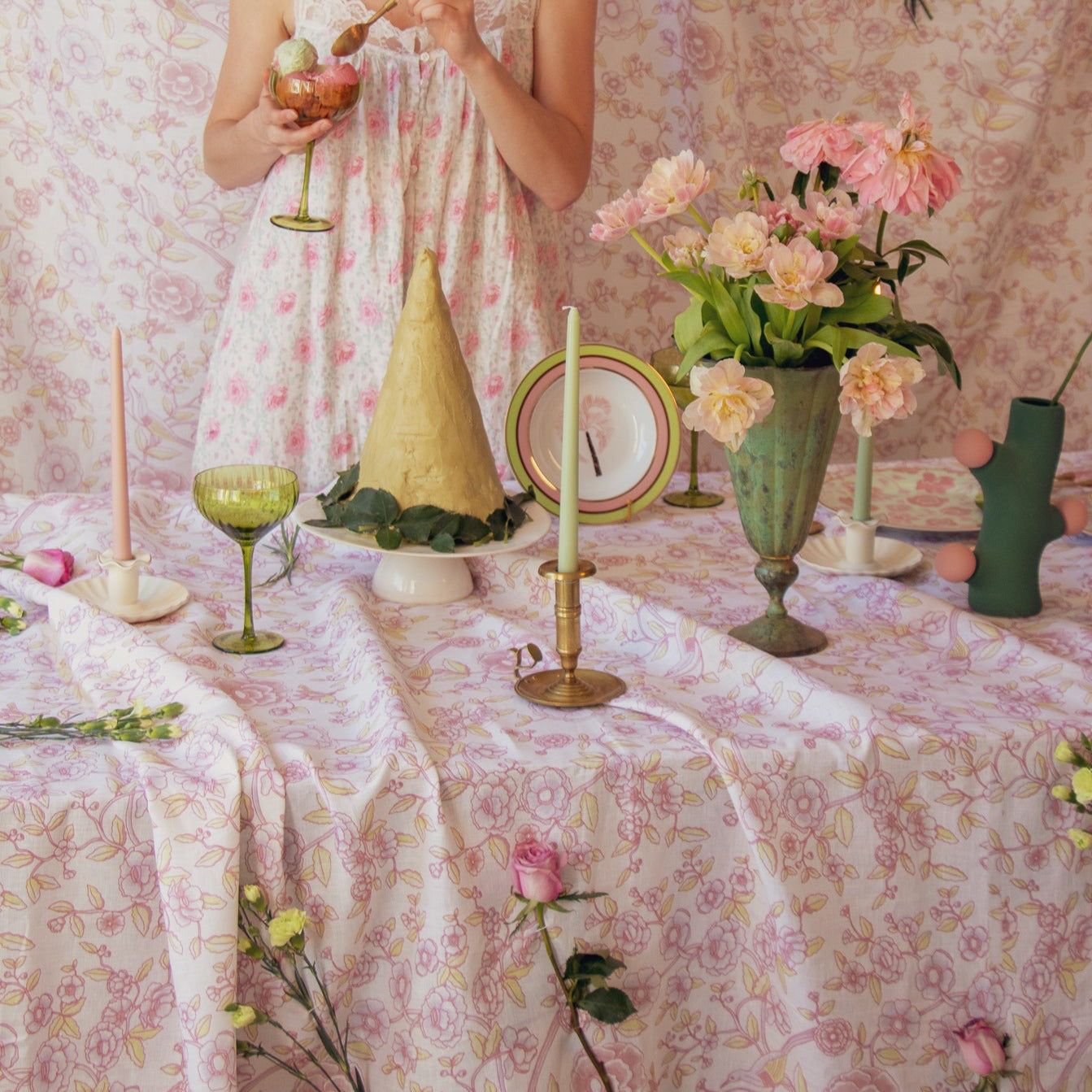 Woman in a floral dress standing behind a table set with decorative items including candles, vases, and flowers.