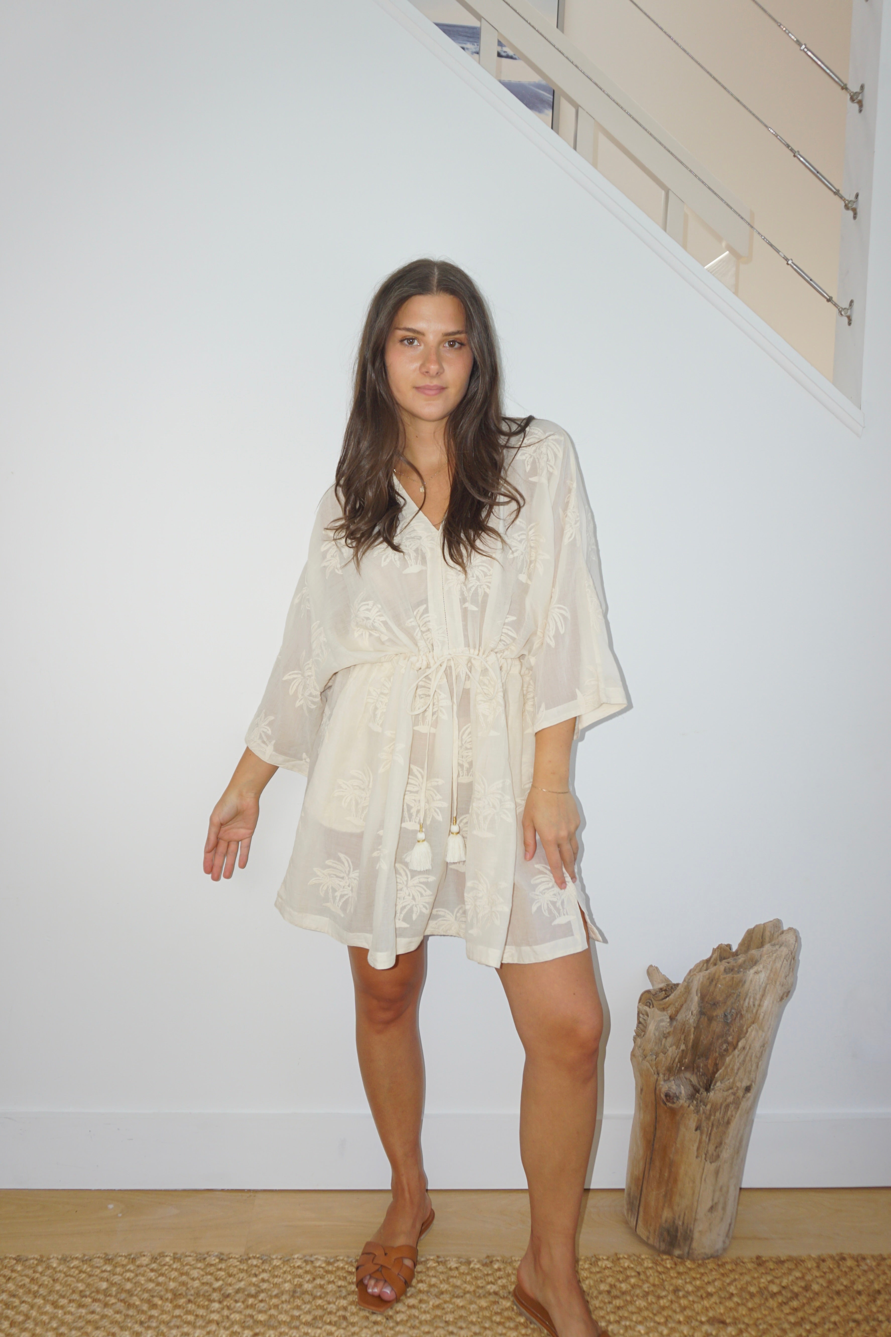 Woman wearing a white dress standing next to a piece of driftwood indoors.