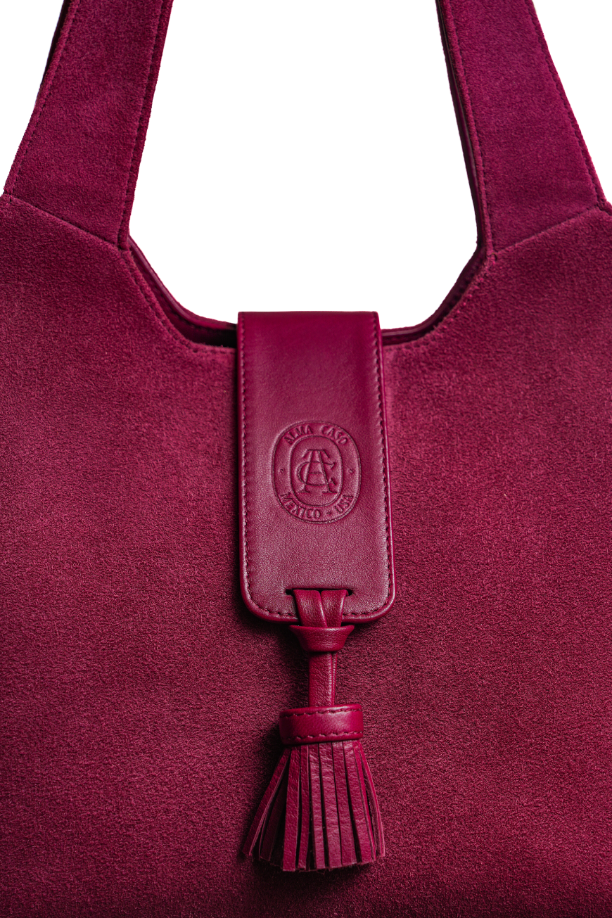 A close-up of a deep magenta suede shoulder bag featuring a matching leather tassel keychain with an embossed circular logo that reads 'MILANO AUTO S.A.