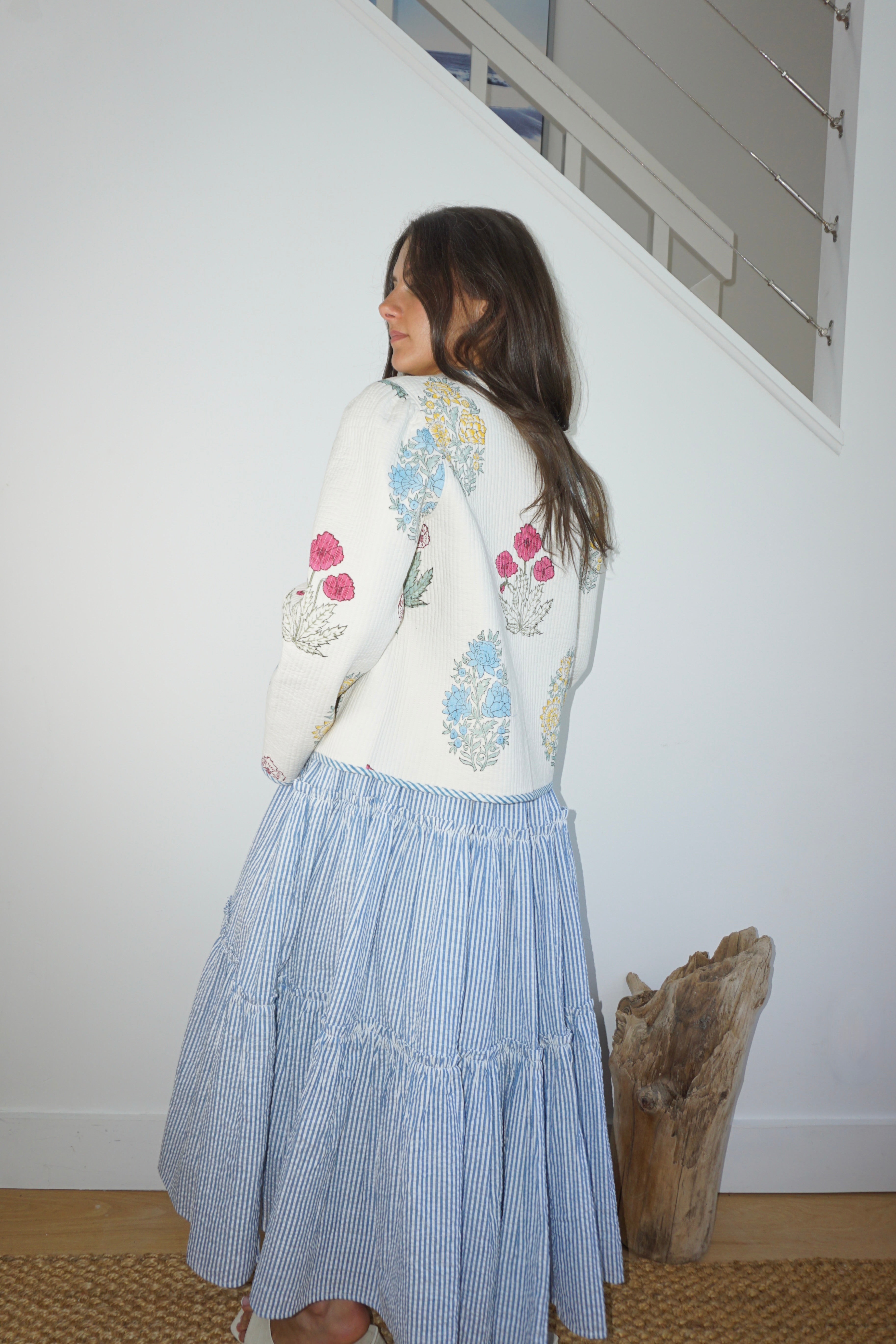 Woman wearing a floral embroidered sweater and blue skirt standing in a room with a white wall and wooden floor.