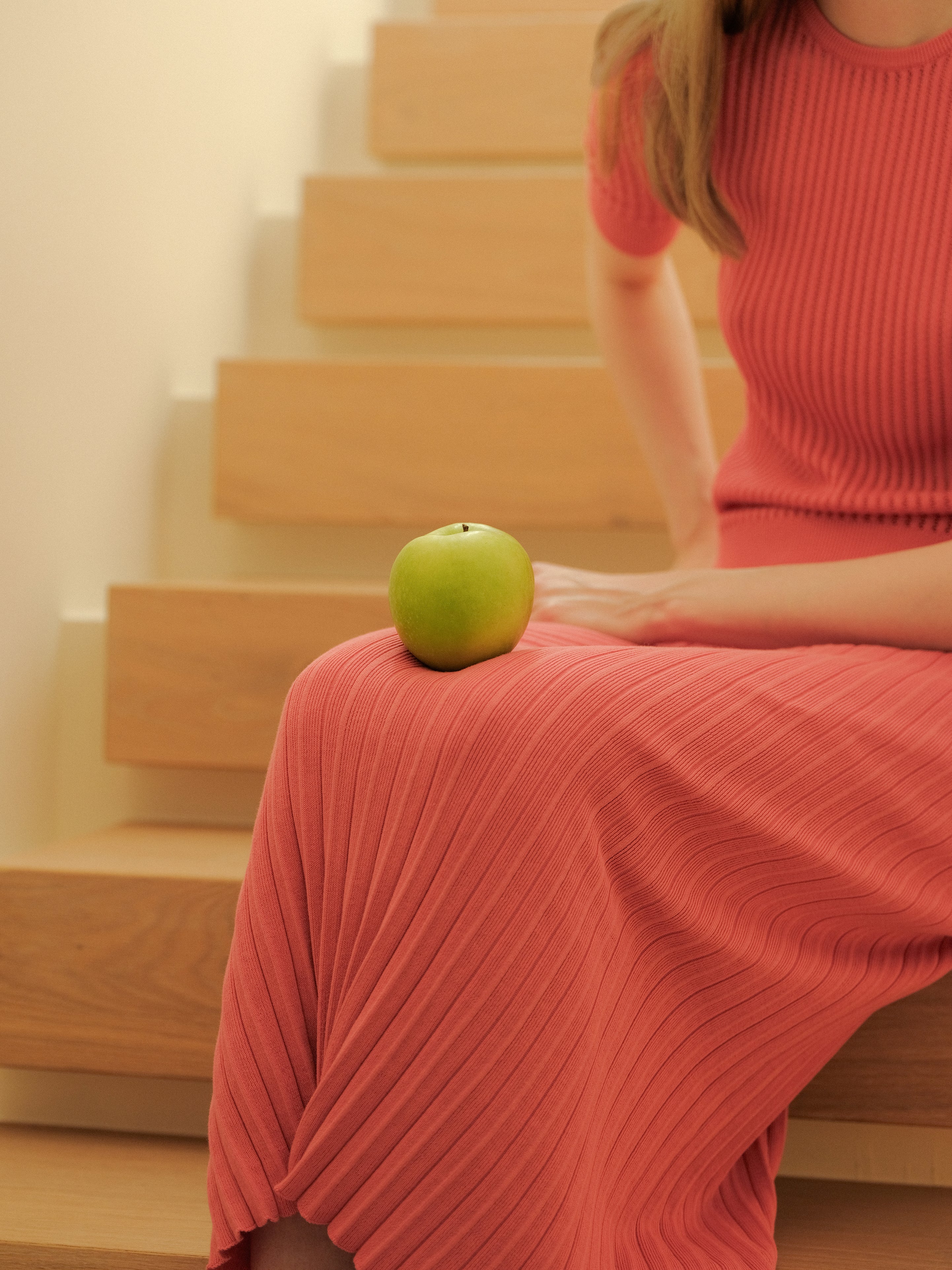 Person in a pink dress sitting on wooden stairs with a green apple on their lap.