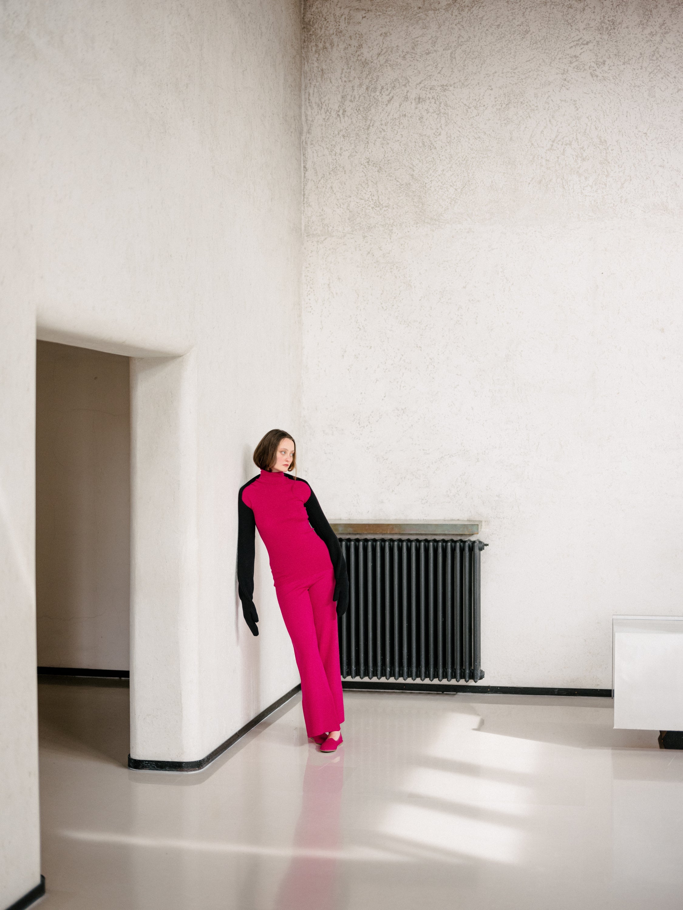 Woman in a bright pink jumpsuit standing in a minimalistic room with white walls and a radiator.