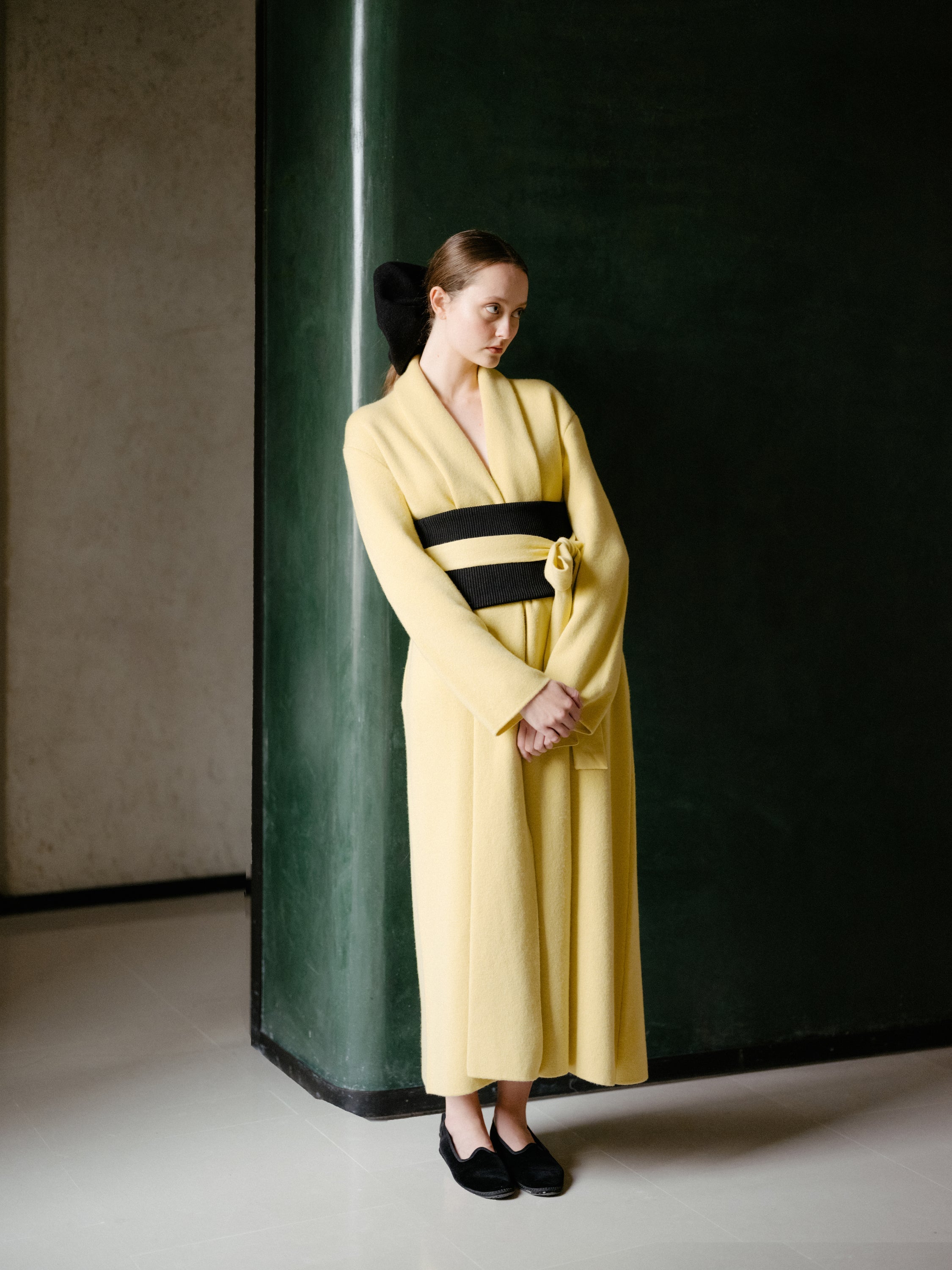 Woman in a long yellow robe with a black belt standing against a dark wall.