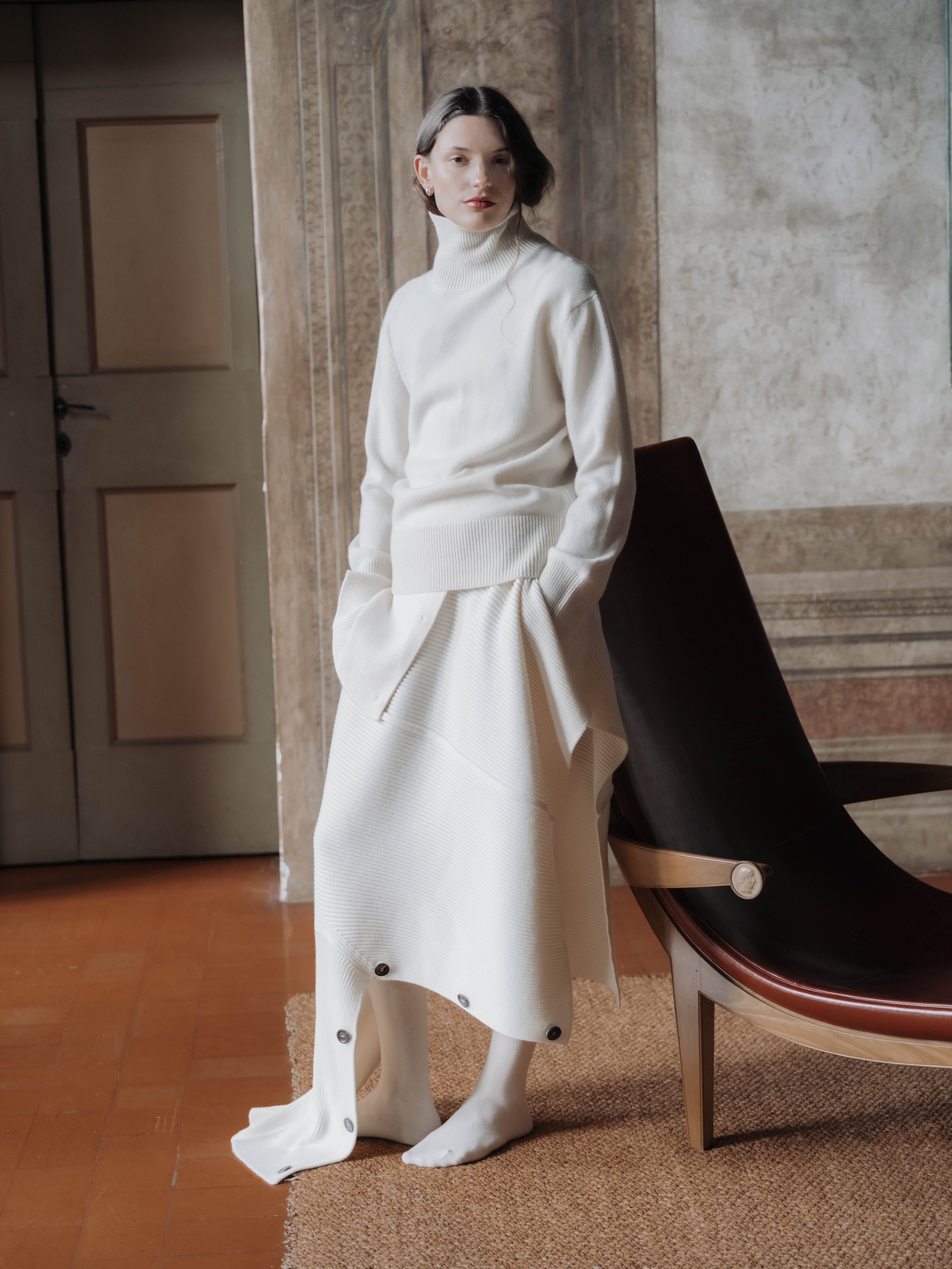 Woman wearing a white turtleneck with a cream-colored cape cardigan as a skirt in an elegant room with a black modern chair.