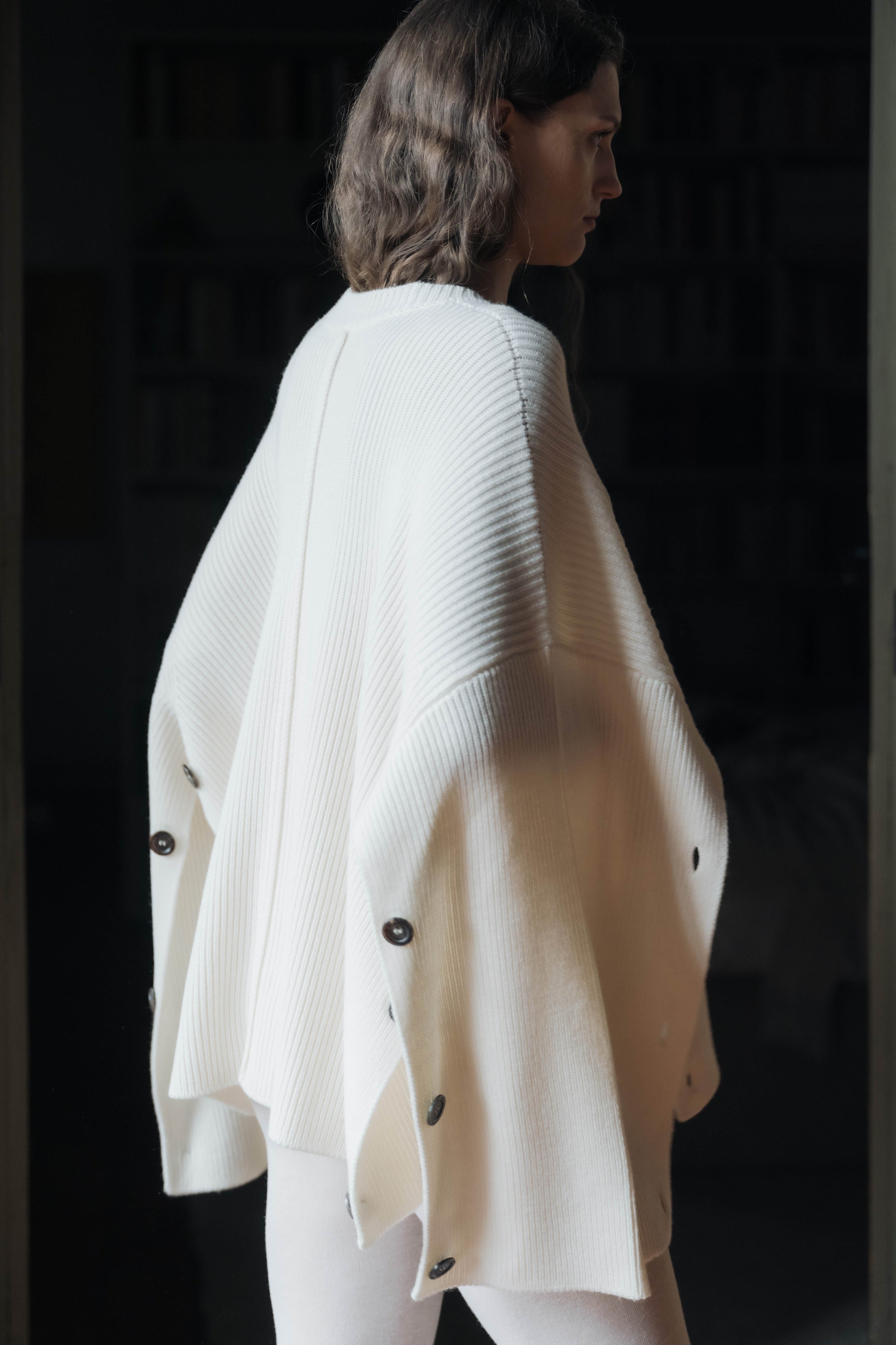 Woman wearing a white knit poncho with black buttons in a dark setting
