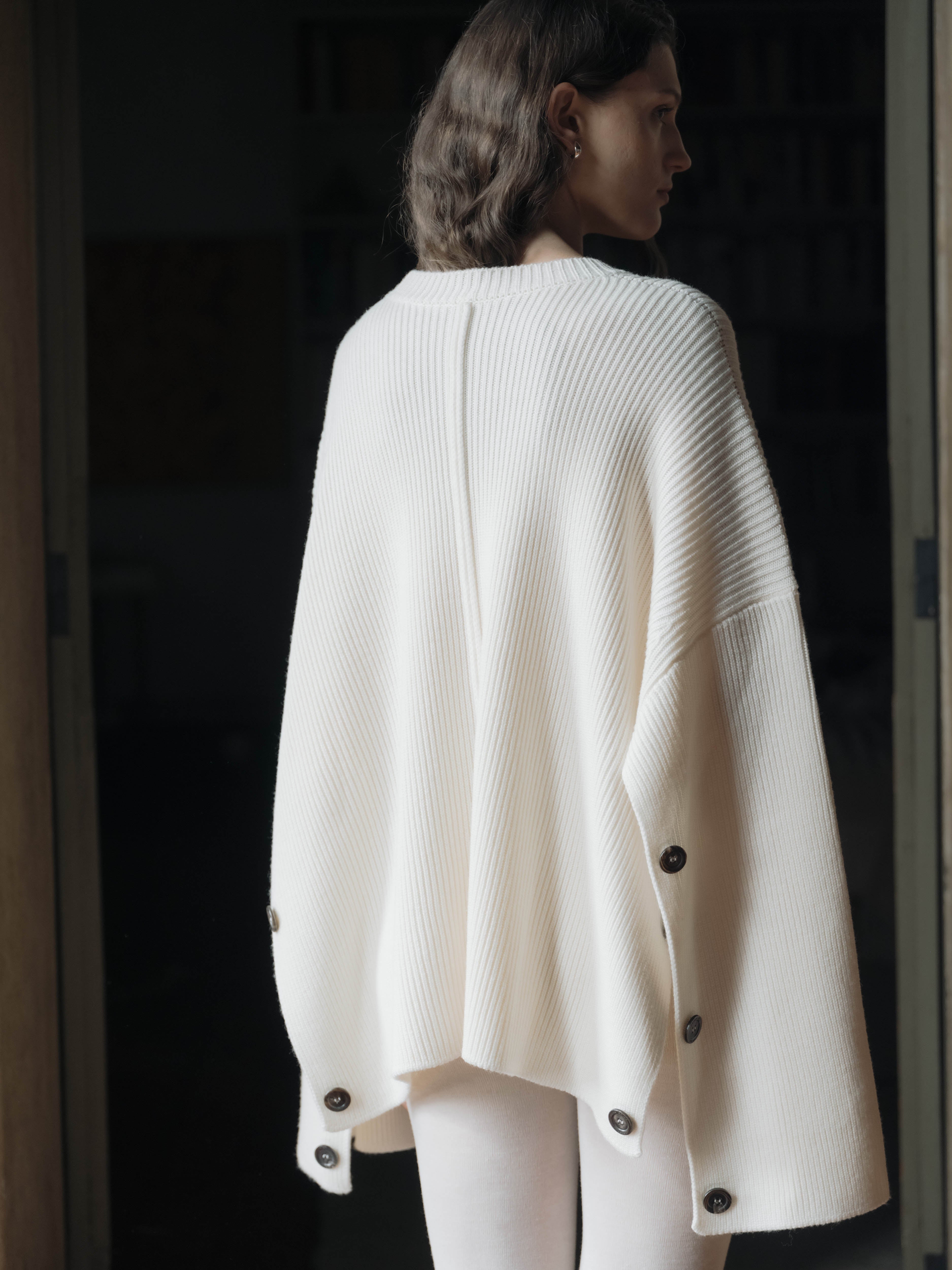 Woman wearing a white knit cape cardigan with dark buttons in a dimly lit room.