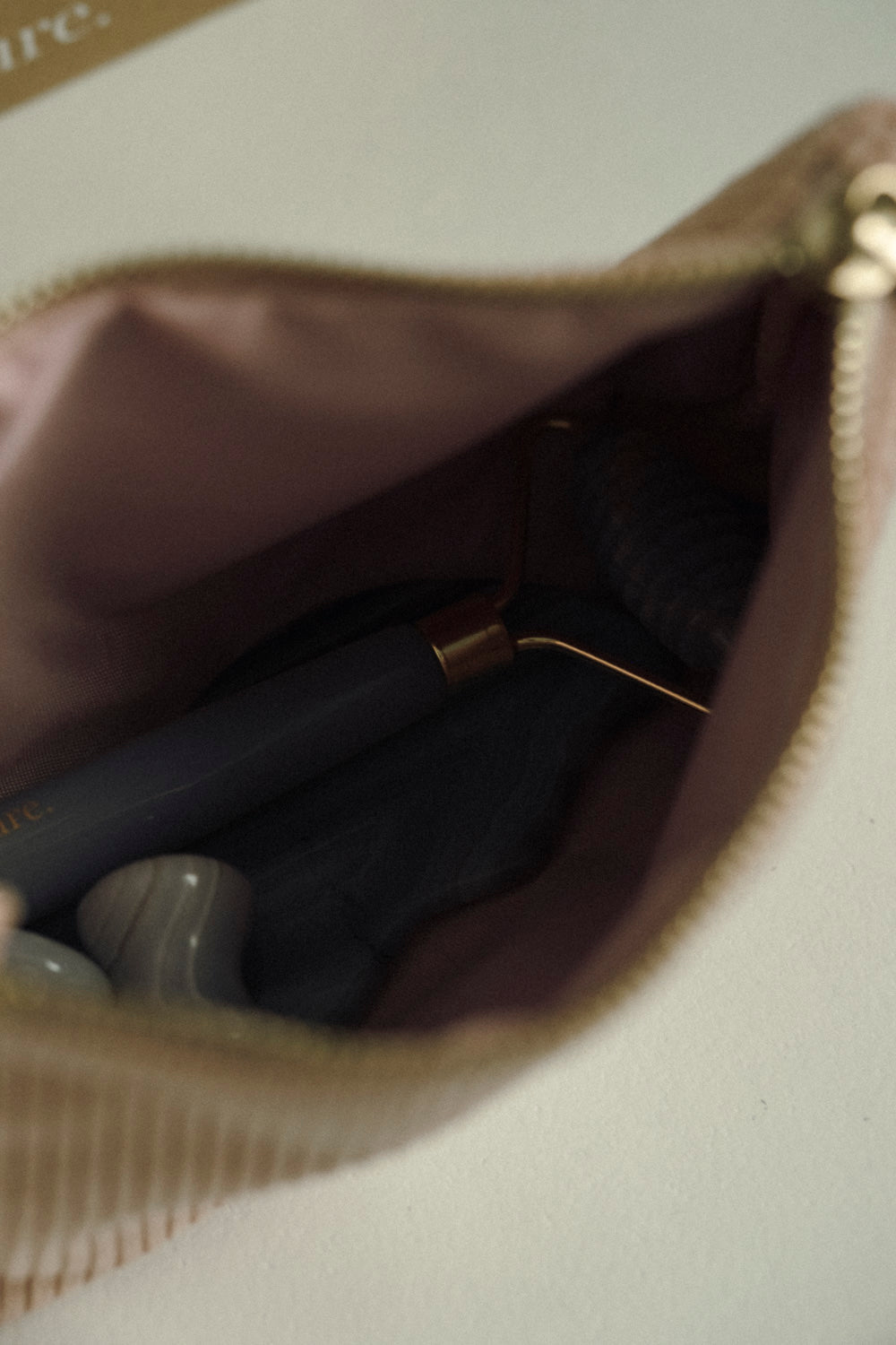 Close-up of a beige zippered pouch with a gold zipper, showing a dark gray cosmetic pen and small white stones inside.