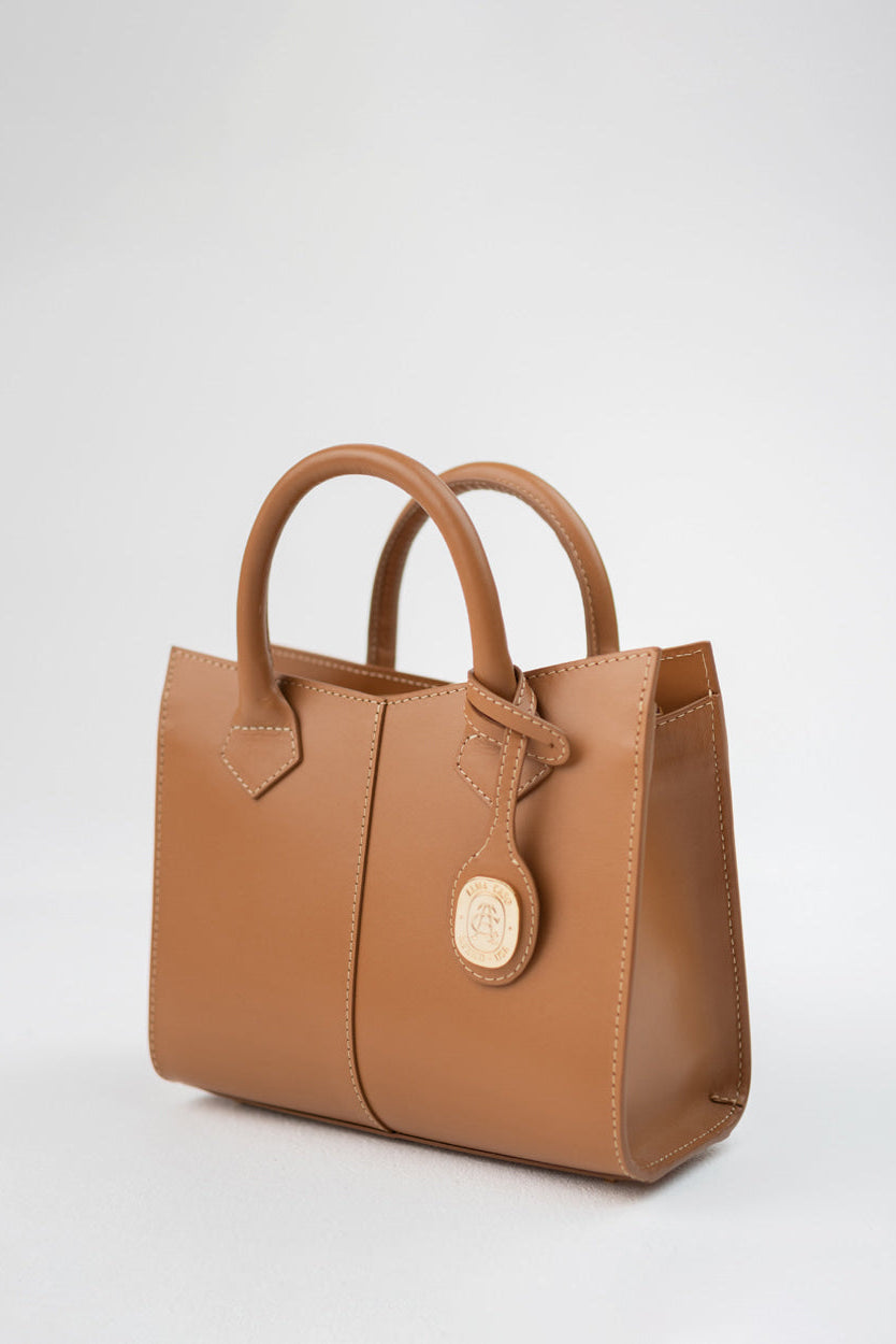 A tan leather handbag with double top handles and a round gold-tone logo medallion, photographed on a clean light gray studio background.