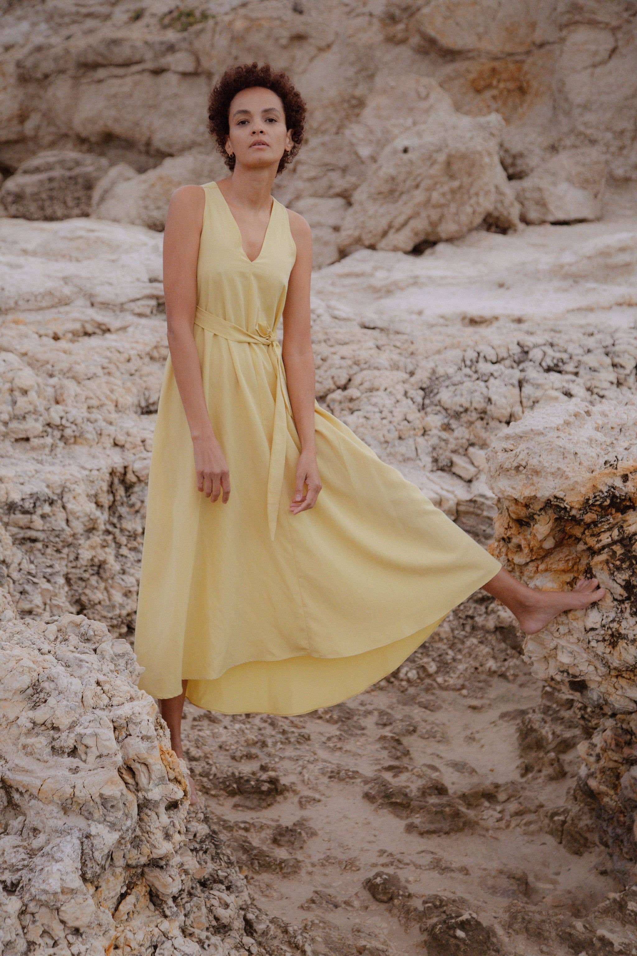 A woman standing on a rocky beach wearing a yellow, long, sleeveless dress with a v-neckline and side seam pockets.