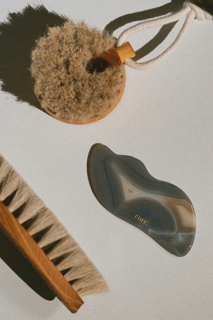 A glossy, lip-shaped massage or beauty tool made of marbled gray agate with the brand name 'cure.' in gold, displayed on a white surface next to natural bristle brushes.