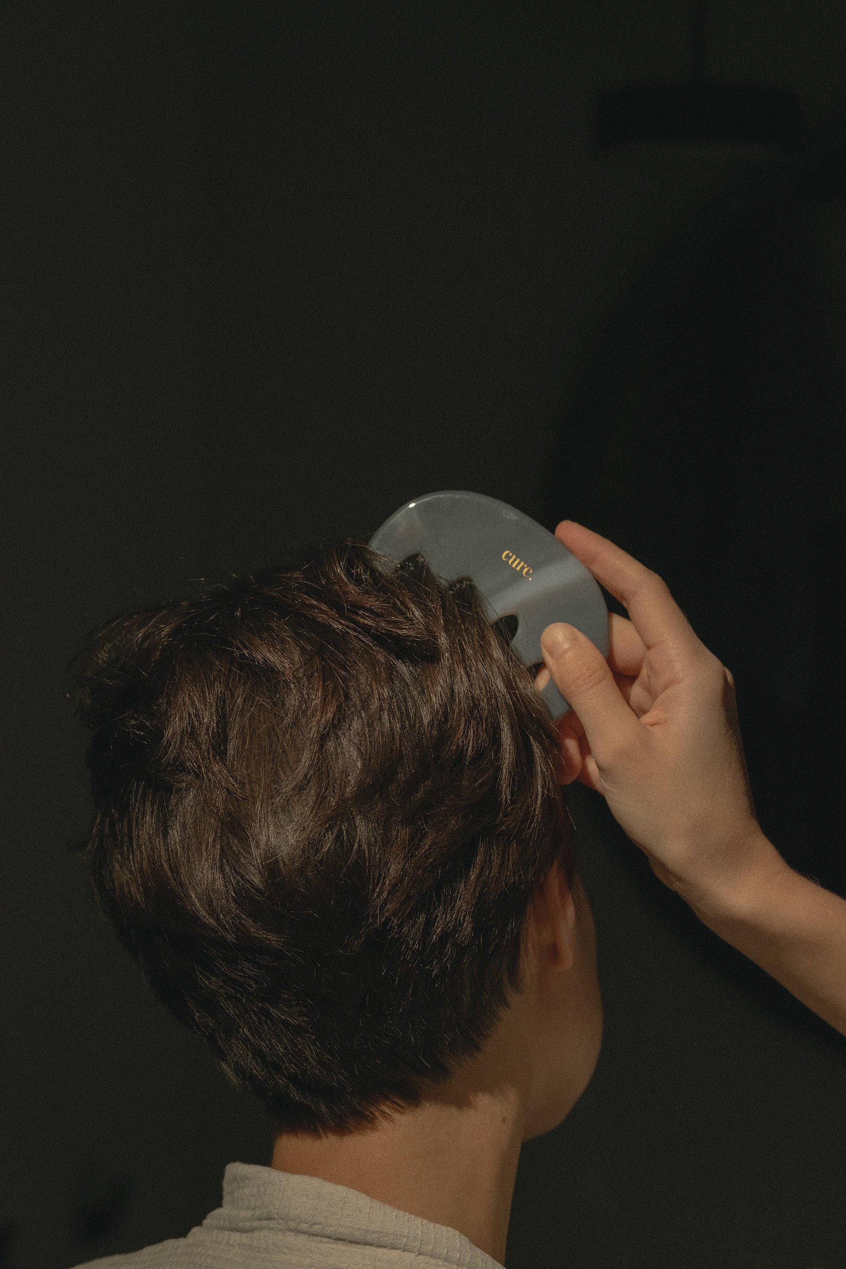A person viewed from behind with short brown hair, having their hair brushed with a gray oval hairbrush held by another person's hand against a dark background.