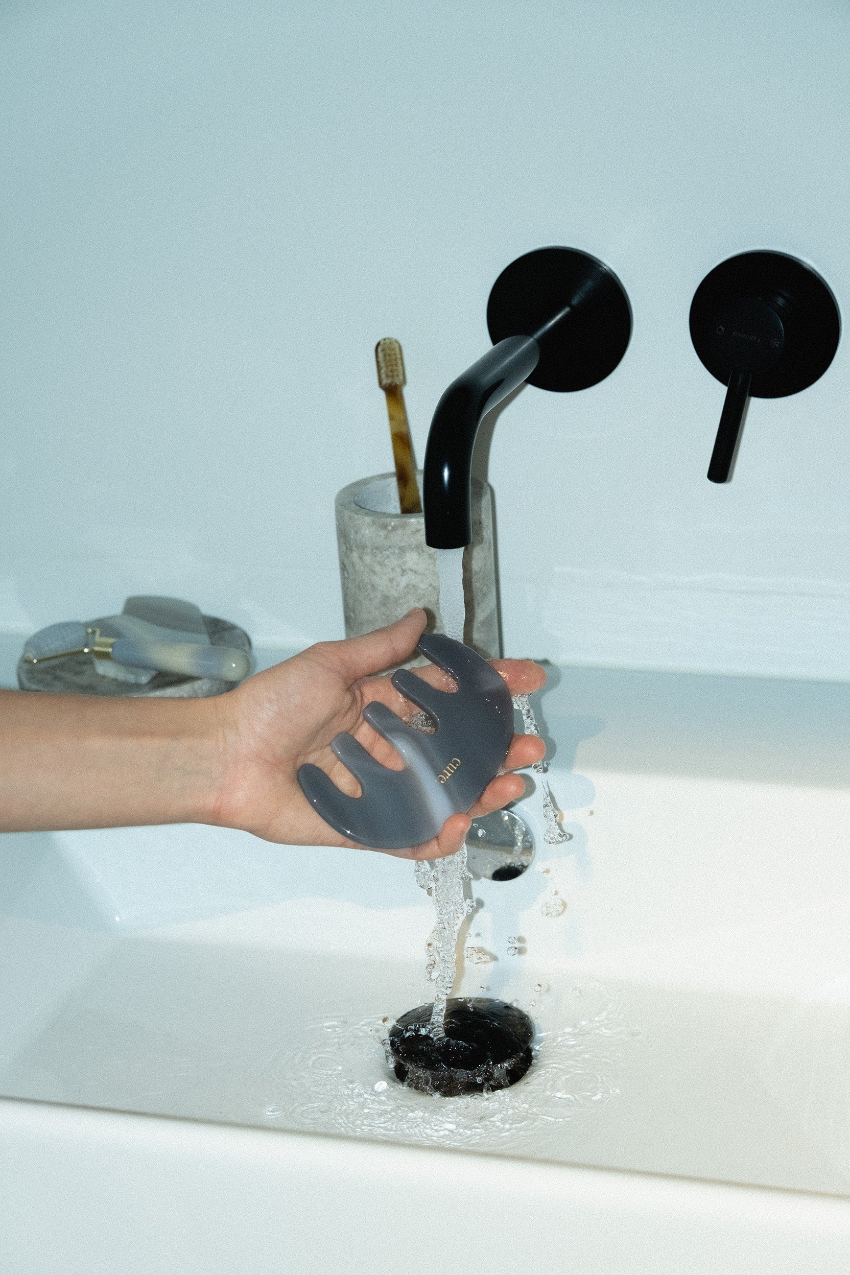 A hand holding a dark gray translucent silicone scrubber under running water from a modern black faucet in a white sink.