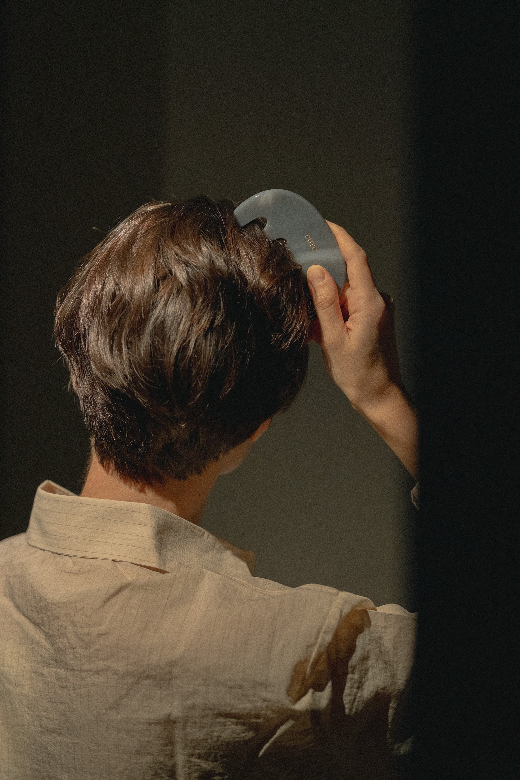 A person viewed from behind wearing a light beige linen shirt and holding a gray hairbrush against their brown hair in a dimly lit setting.