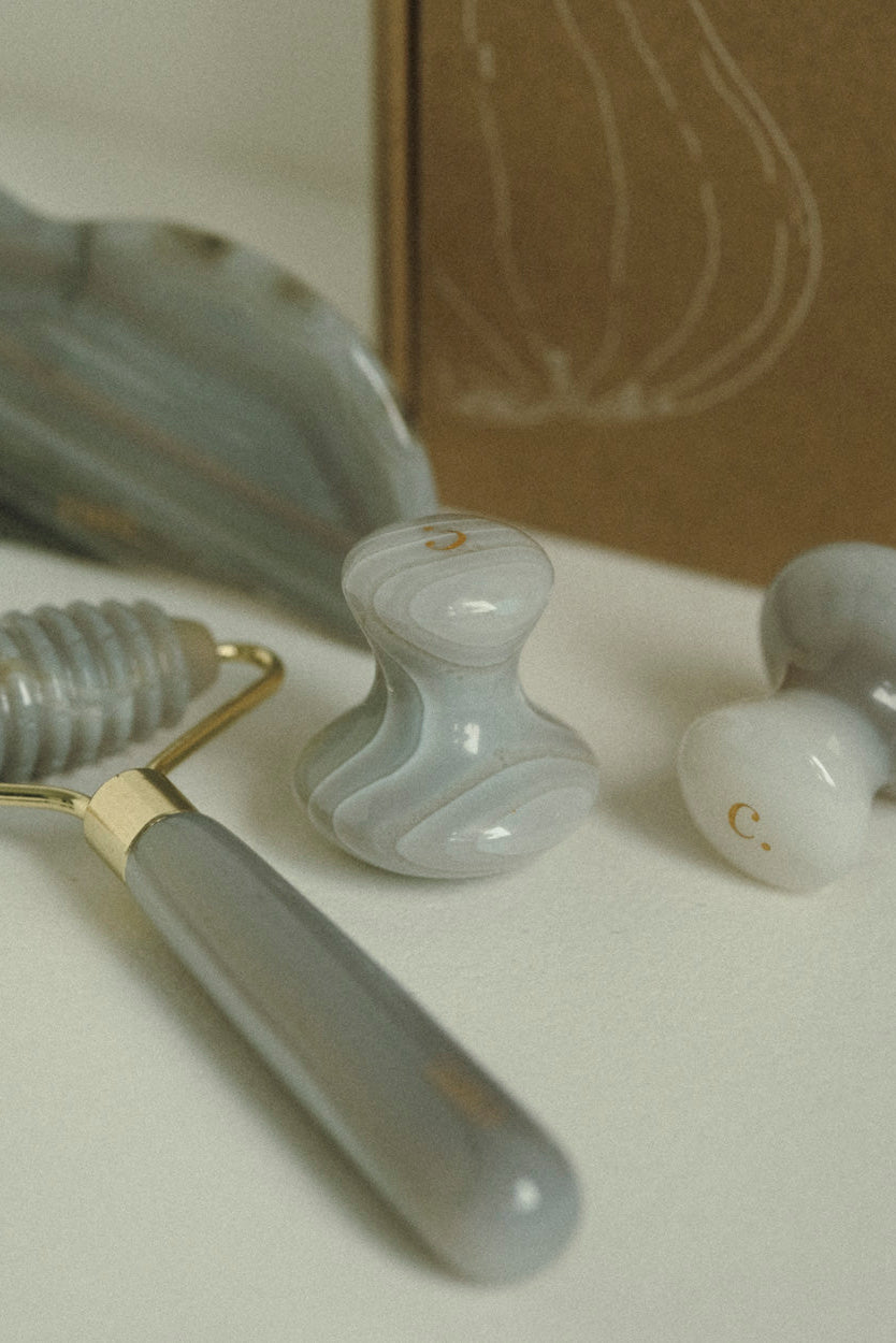 A close-up of a facial massage set featuring a gray marbled stone roller, two matching massage heads, and a brown cardboard box in the background.