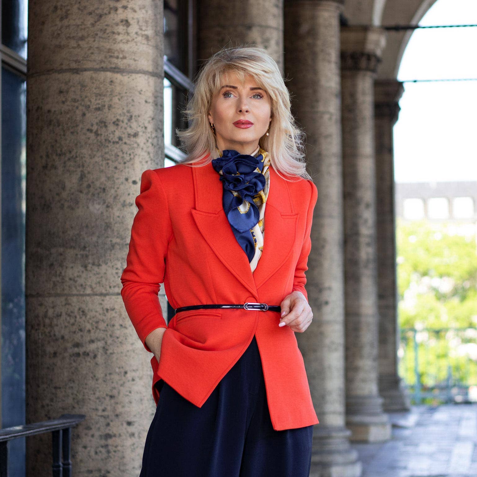 Woman in a red coat with a blue scarf standing in front of stone columns.