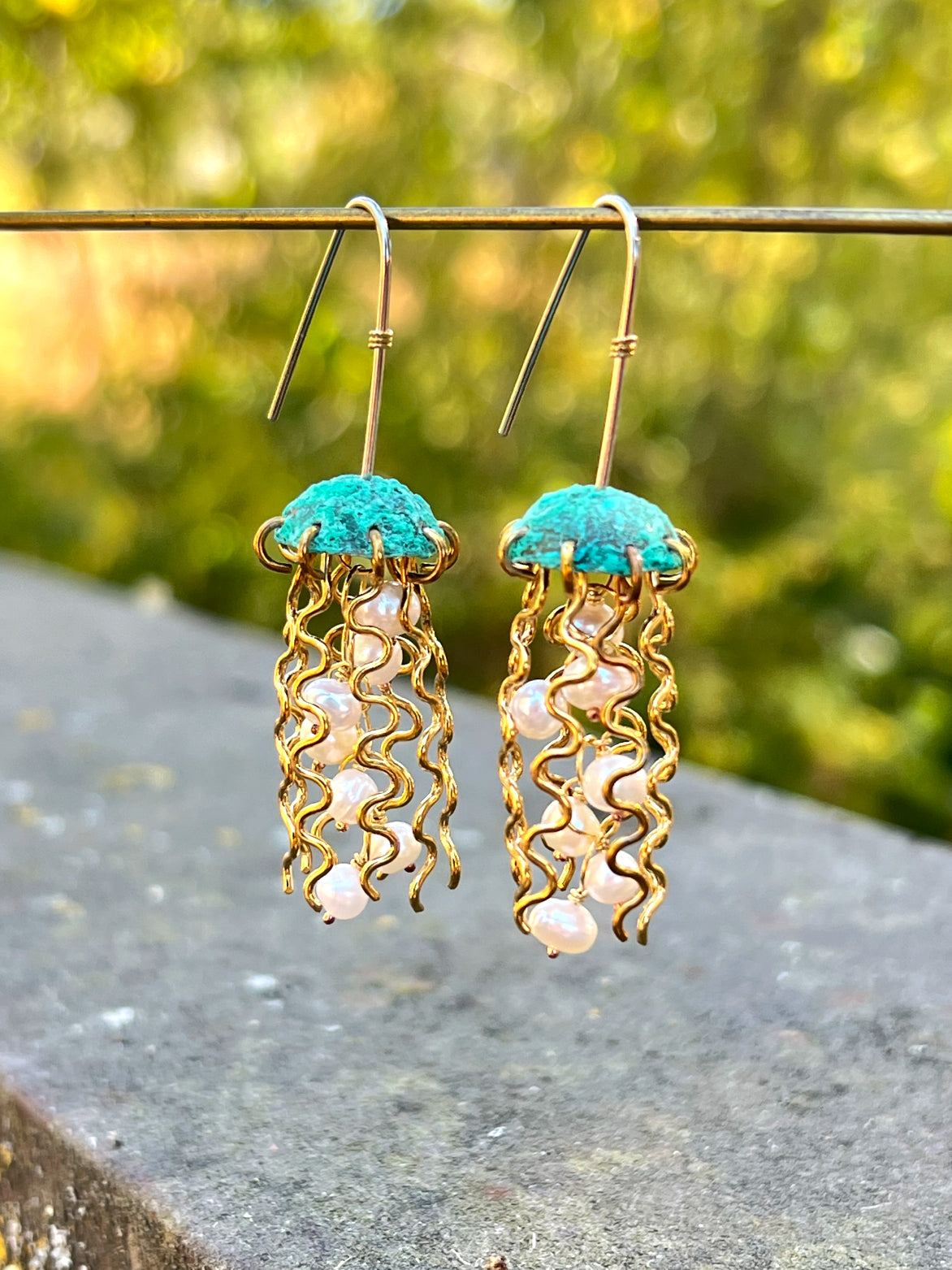 Jellyfish-shaped earrings with turquoise tops and gold wavy designs on a blurred natural background