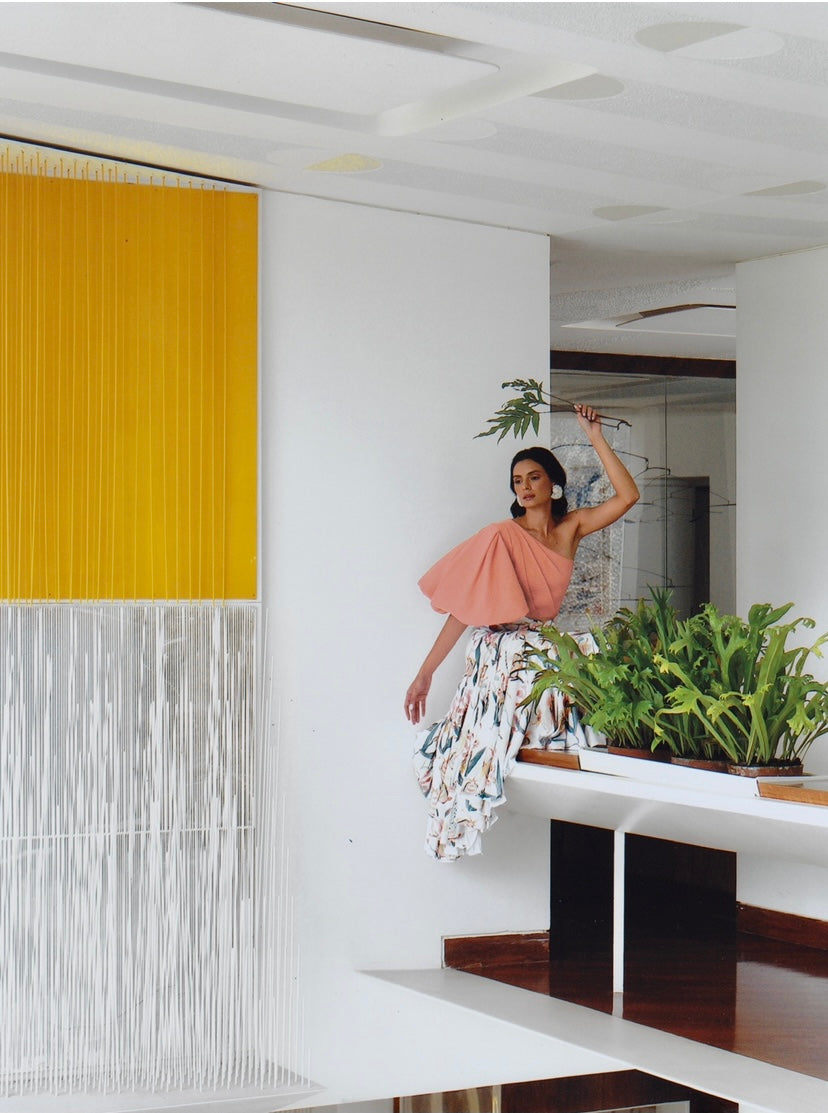 Woman wearing a peach colored one-sleeve top and standing in a modern interior with a yellow accent wall and plants.