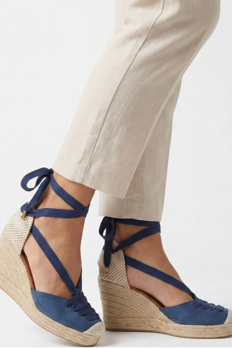 Blue wedge sandals with espadrille sole worn with beige pants on a white background
