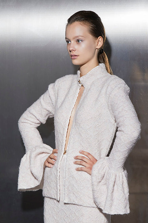 A cream colored, peplum, knitted blazer with bell sleeves on a woman standing with her hands on her hips.