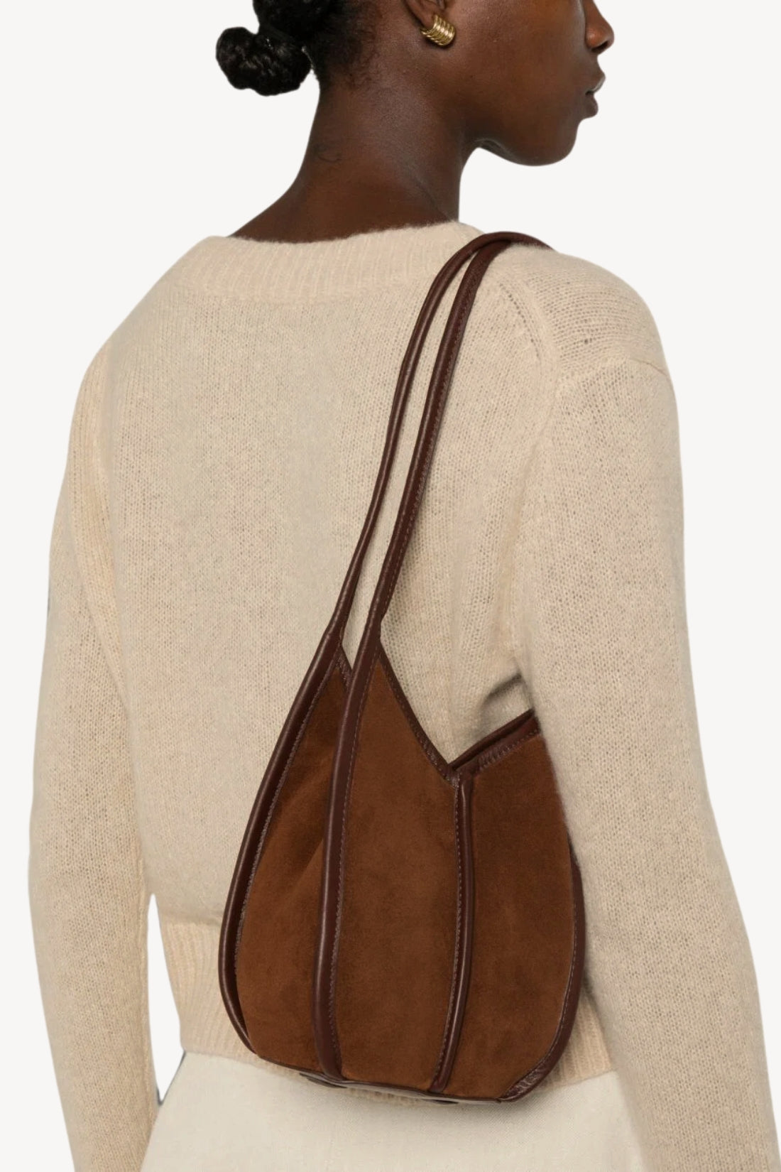 A woman wearing a cream knit sweater is shown from the back, carrying a brown suede shoulder bag with dark brown leather trim and straps.