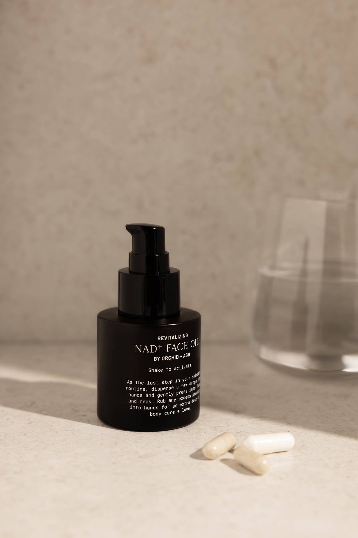 A black bottle of REVITALIZING NAD+ FACE OIL with a black pump, set on a light beige stone surface next to two white capsules, with a blurred glass of water in the background.