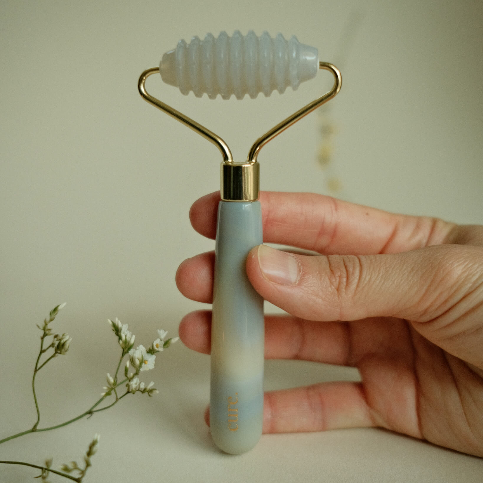 A hand holding a jade roller tool with a ribbed white jade head and a pale blue gradient handle with gold metal accents, set against a soft beige background with small white flowers in the corner.