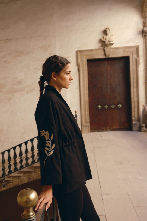 A person standing in a room with classic decor, wearing a long sleeve black jacket with embroidered olive branches on the sleeves.