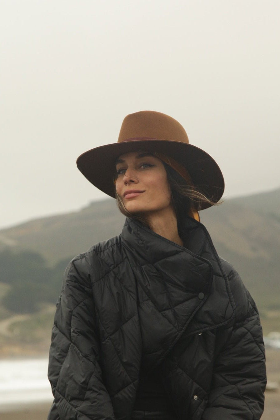 Person wearing a brown hat and black coat standing in a mountainous landscape.