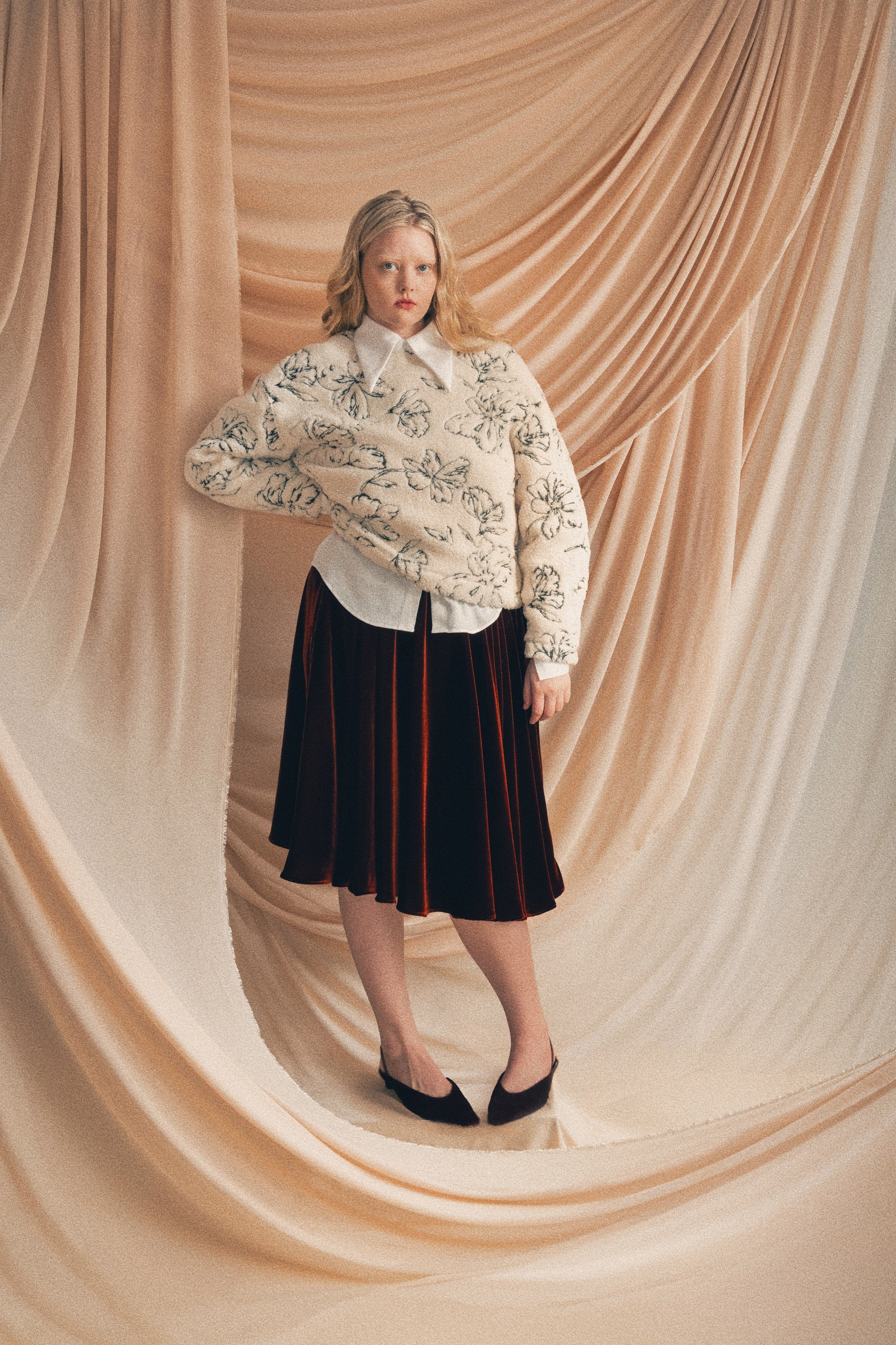 Woman wearing a white blouse with floral patterns and a dark copper red velvet skirt against a beige draped curtain background.