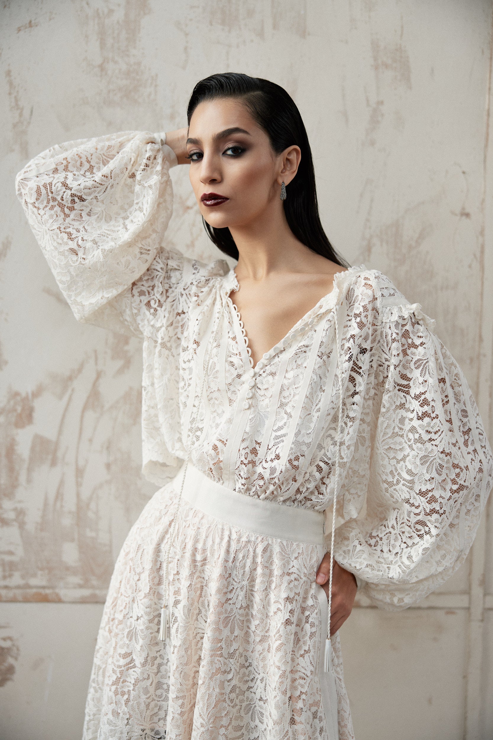 Woman wearing a white lace dress against a textured wall.
