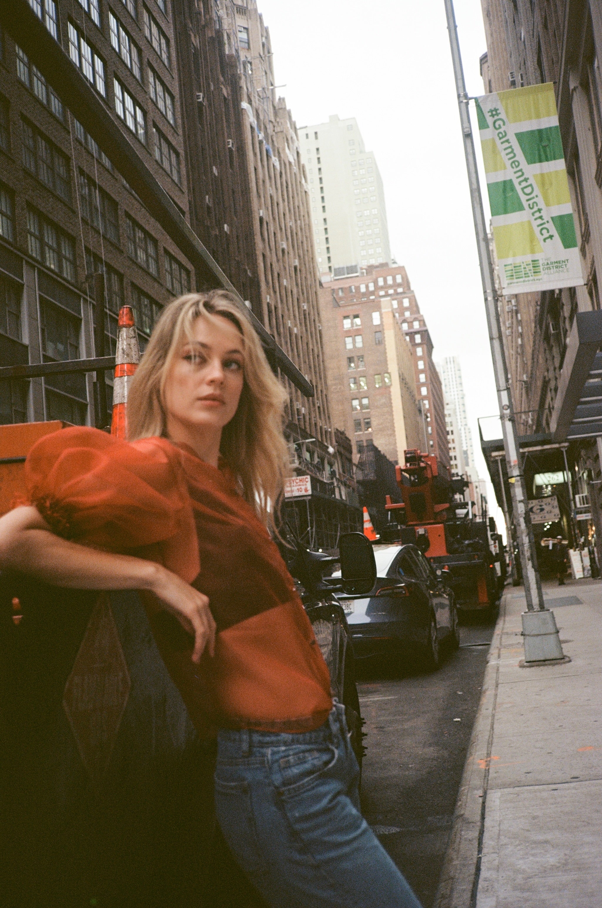 Woman in an orange top and blue jeans standing on a city street with tall buildings in the background.