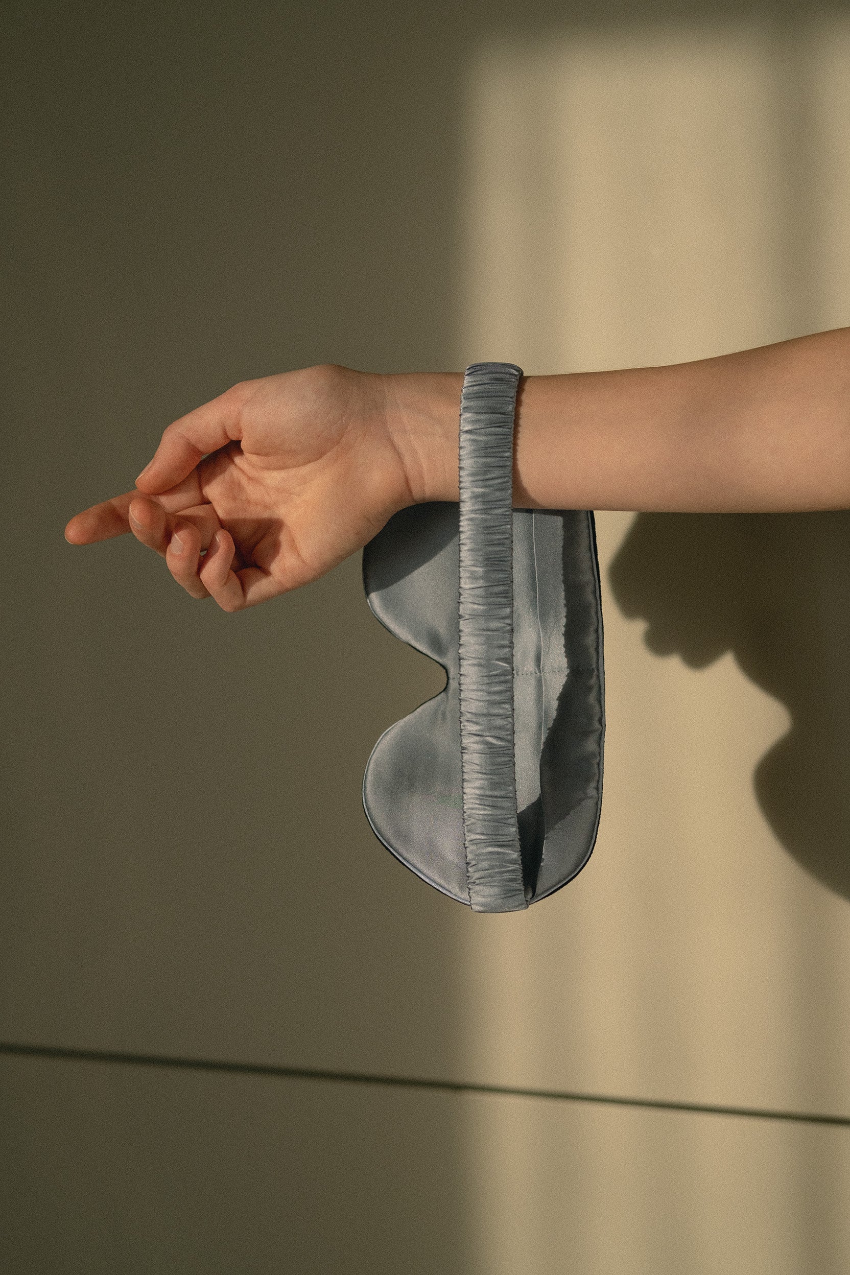 A blue gray sleep mask with a textured, ribbed elastic strap, shown being worn on a person's wrist against a warm-toned beige background with soft window shadows.