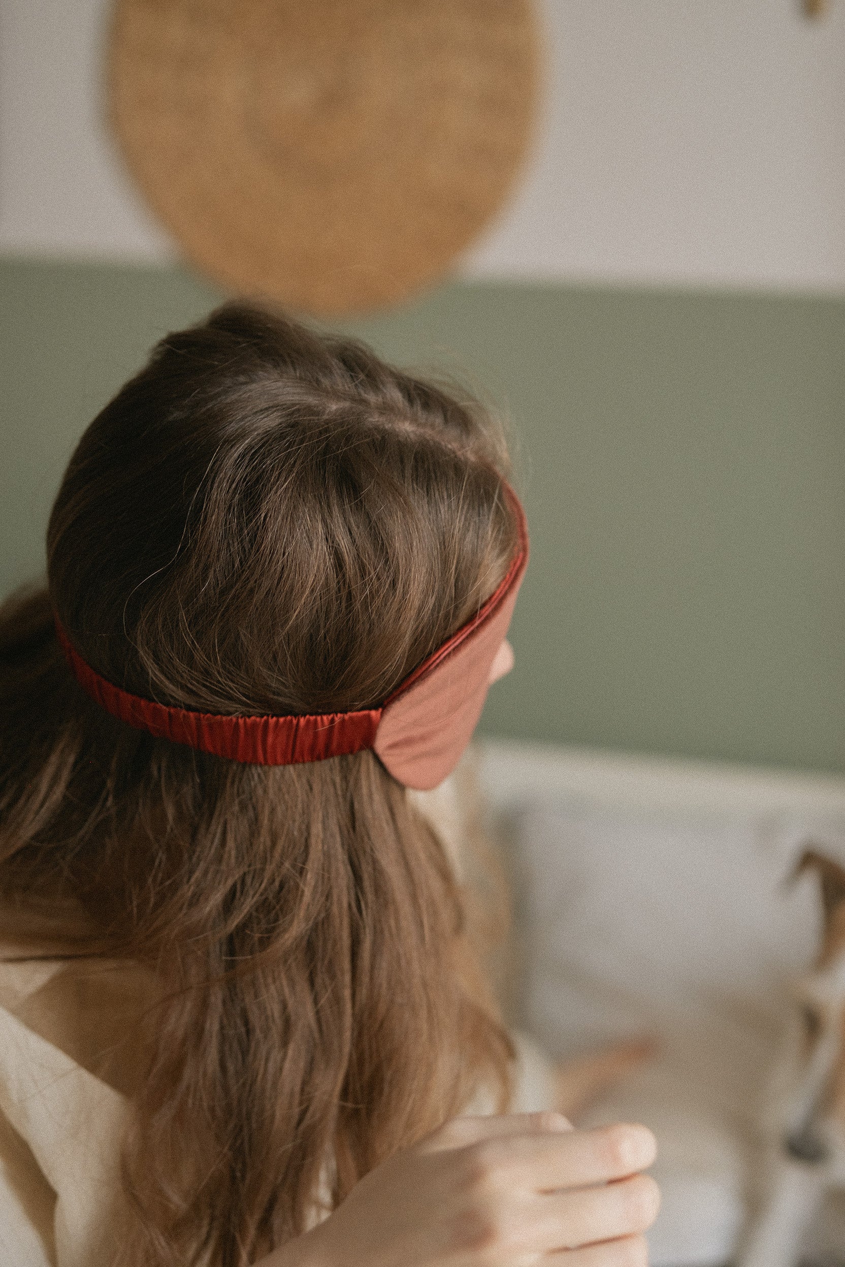 A woman with long brown hair seen from behind, wearing a red velvet eyemask, in a softly lit indoor setting.