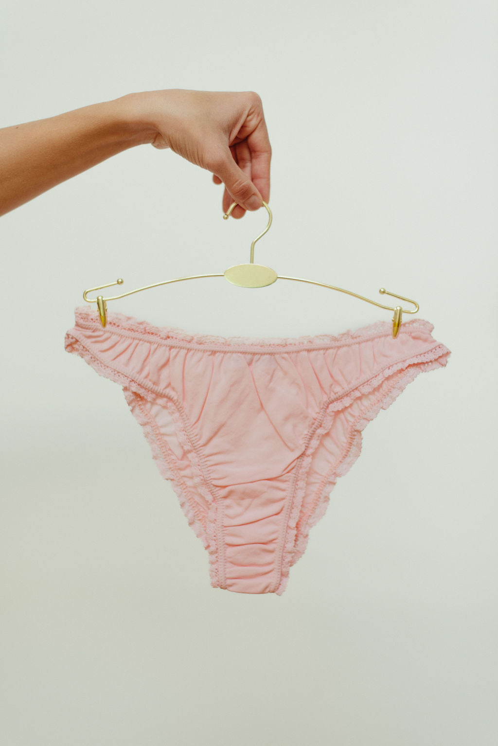 Pink textured underwear on a gold hanger held by a hand against a light background