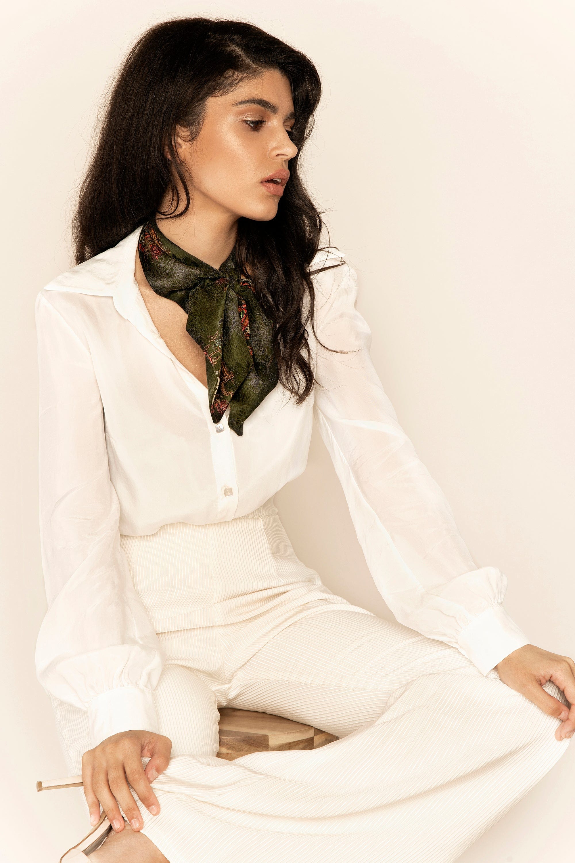 Woman wearing a white outfit with a green scarf, sitting against a plain background