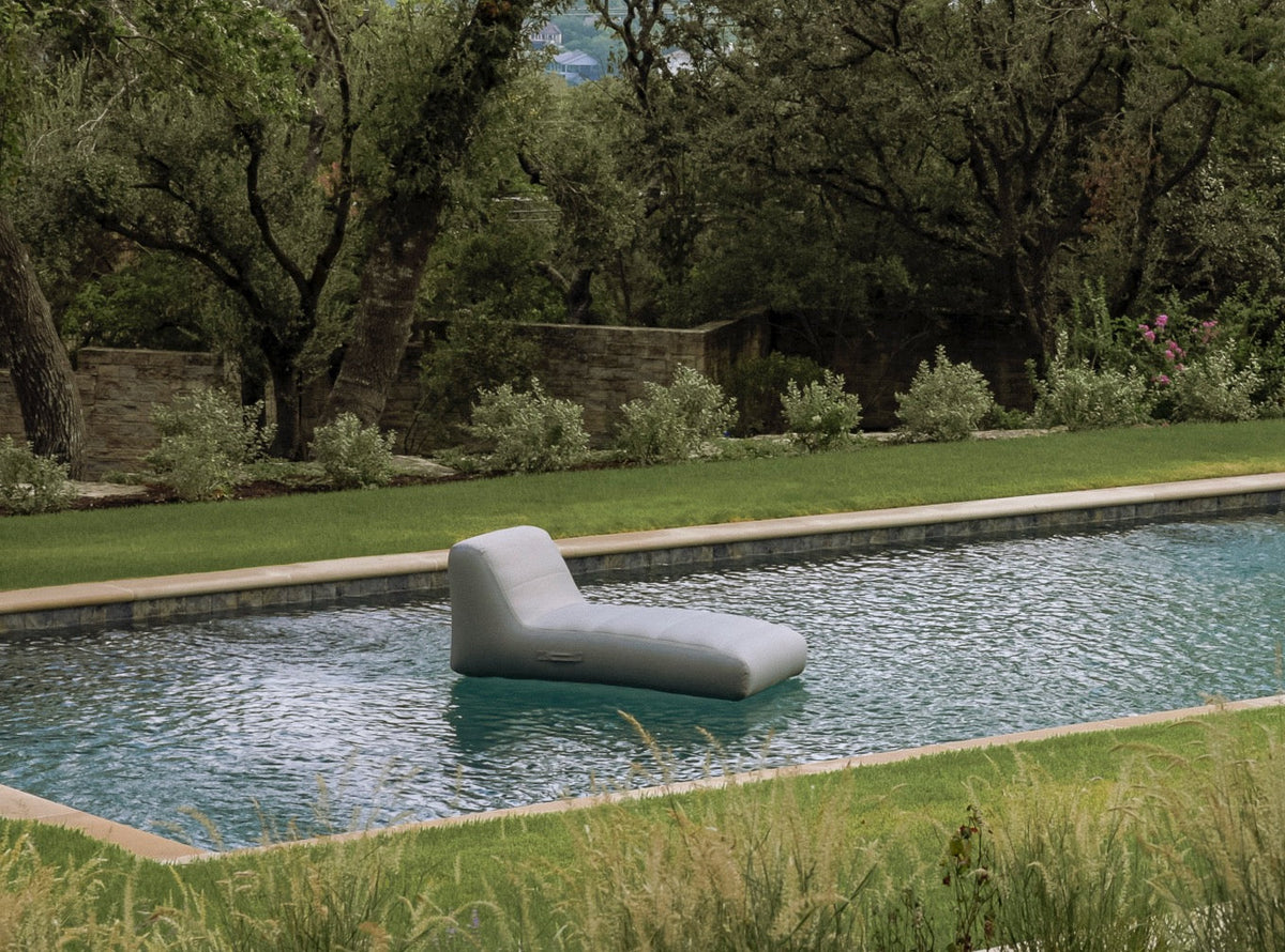 A modern, minimalist light gray chaise lounge floating in a swimming pool, set against a luxurious backyard backdrop with a manicured lawn, stone wall, and lush green trees.