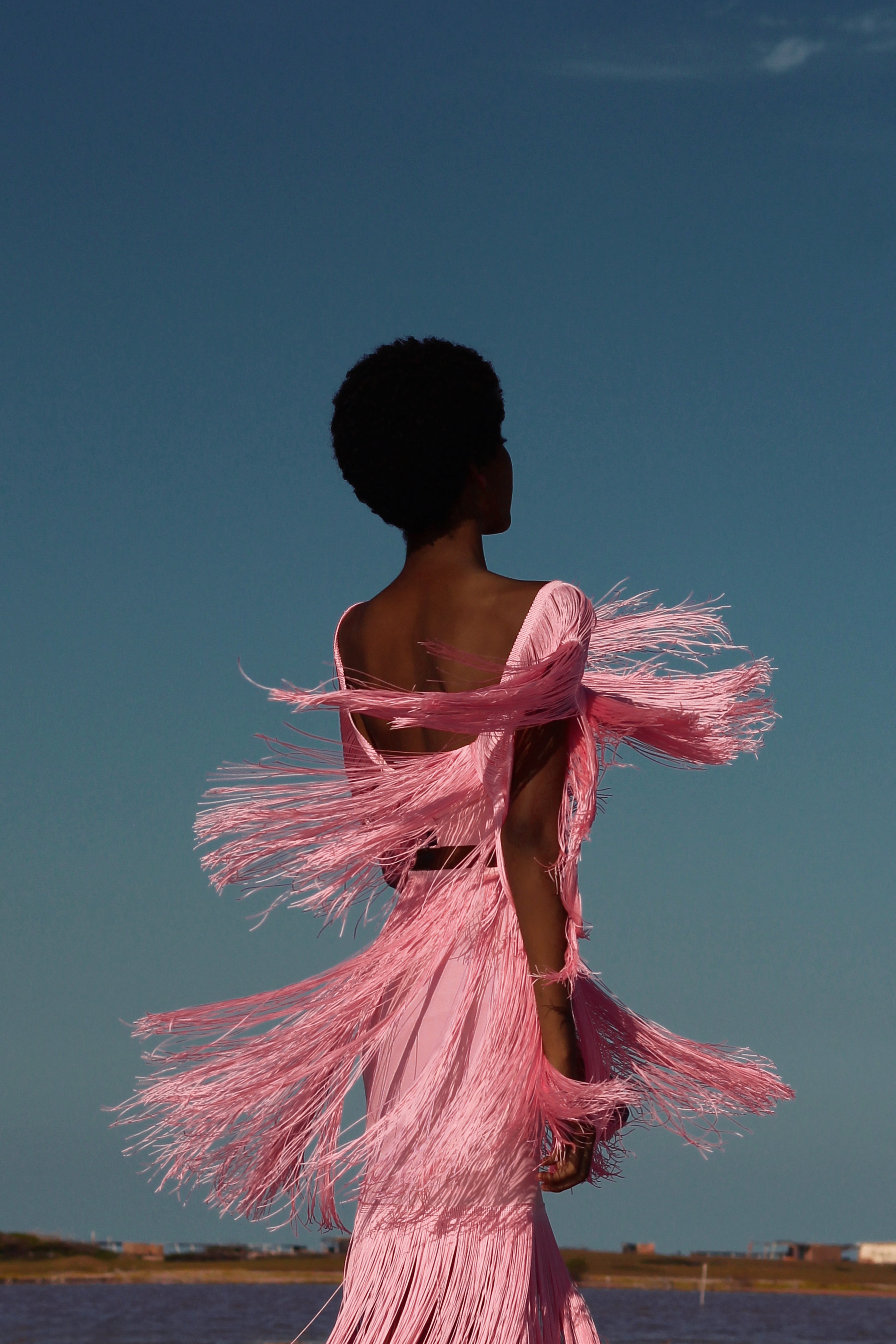 Person wearing a pink fringed dress against a clear blue sky.