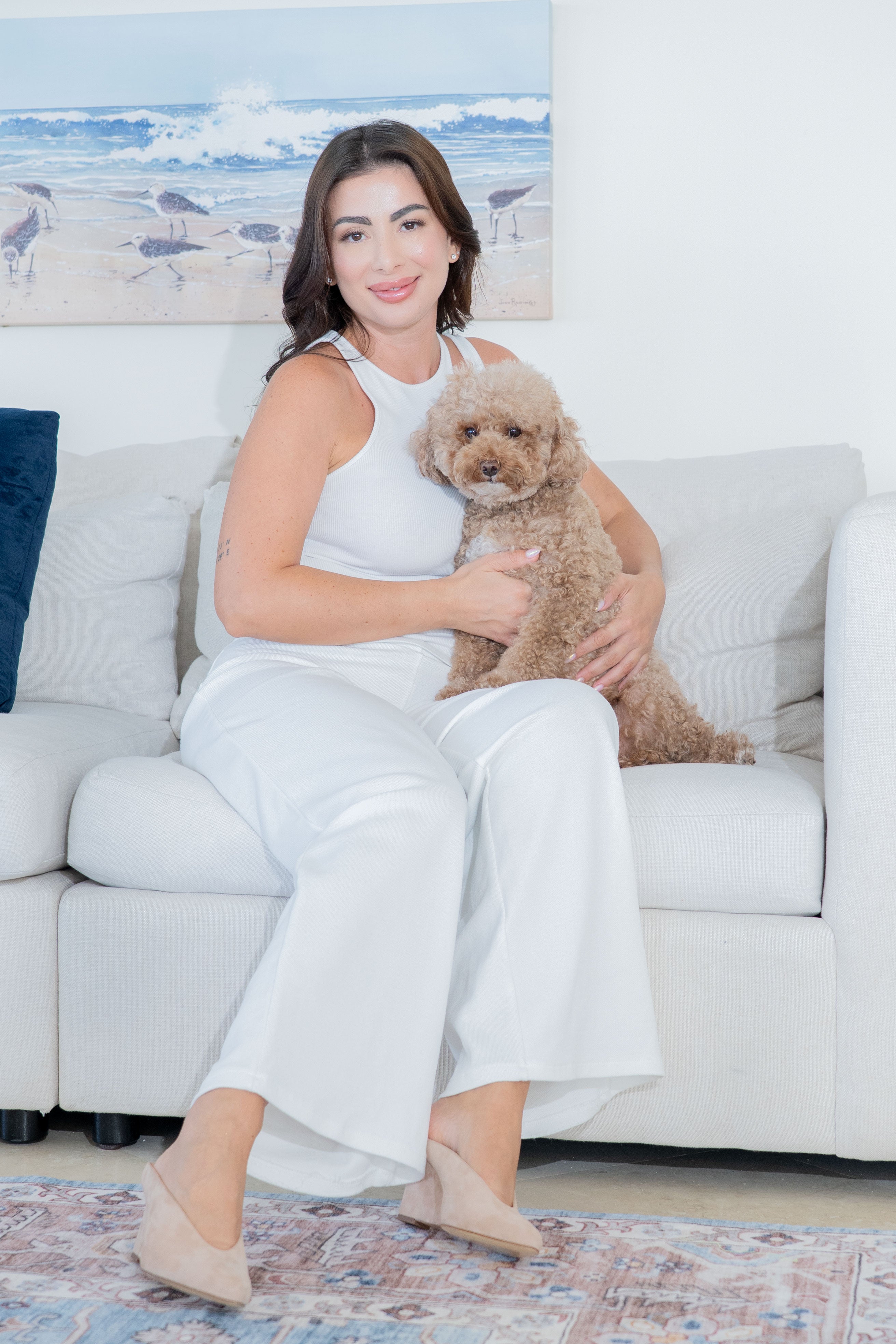 A woman in a white sleeveless top, white wide-leg pants, and suede beige wedged heels sits on a white sofa holding a small fluffy tan dog. The room has a light wall with a beach painting above the sofa, and a patterned rug is visible on the floor.