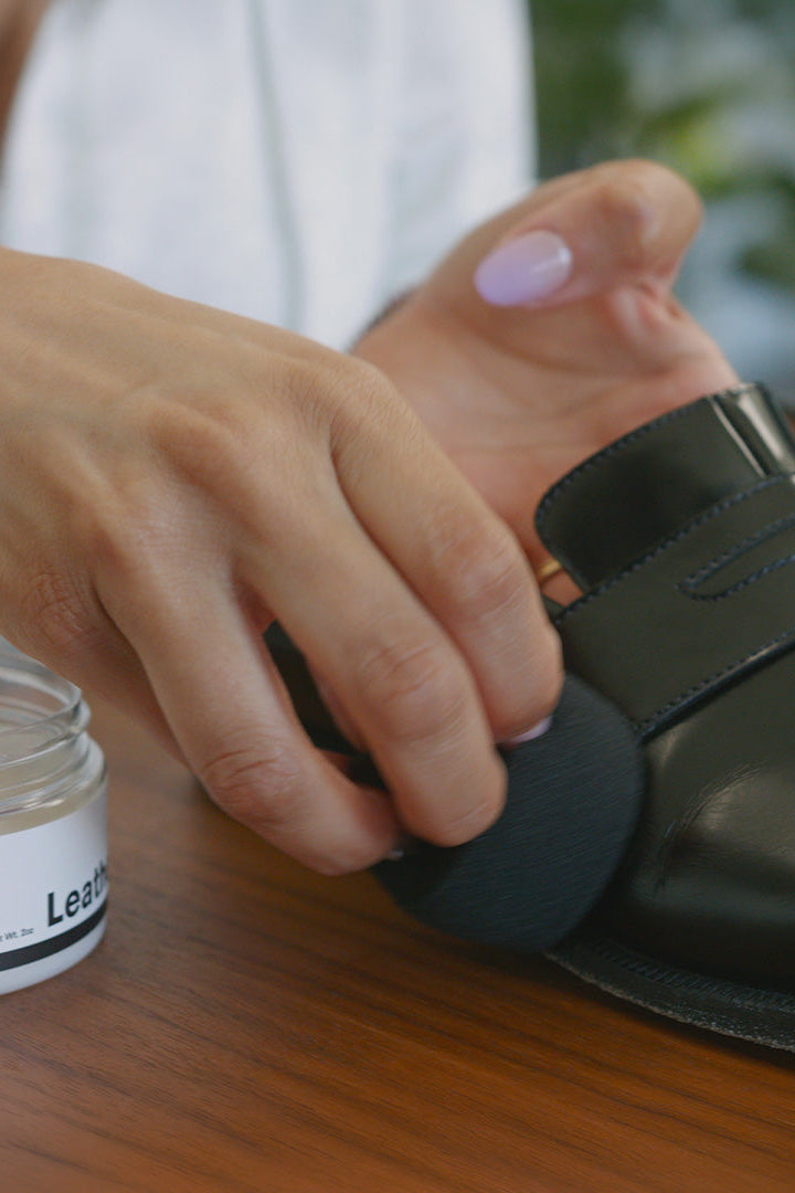 Person applying Jason Markk leather balm to a black shoe on a wooden surface.