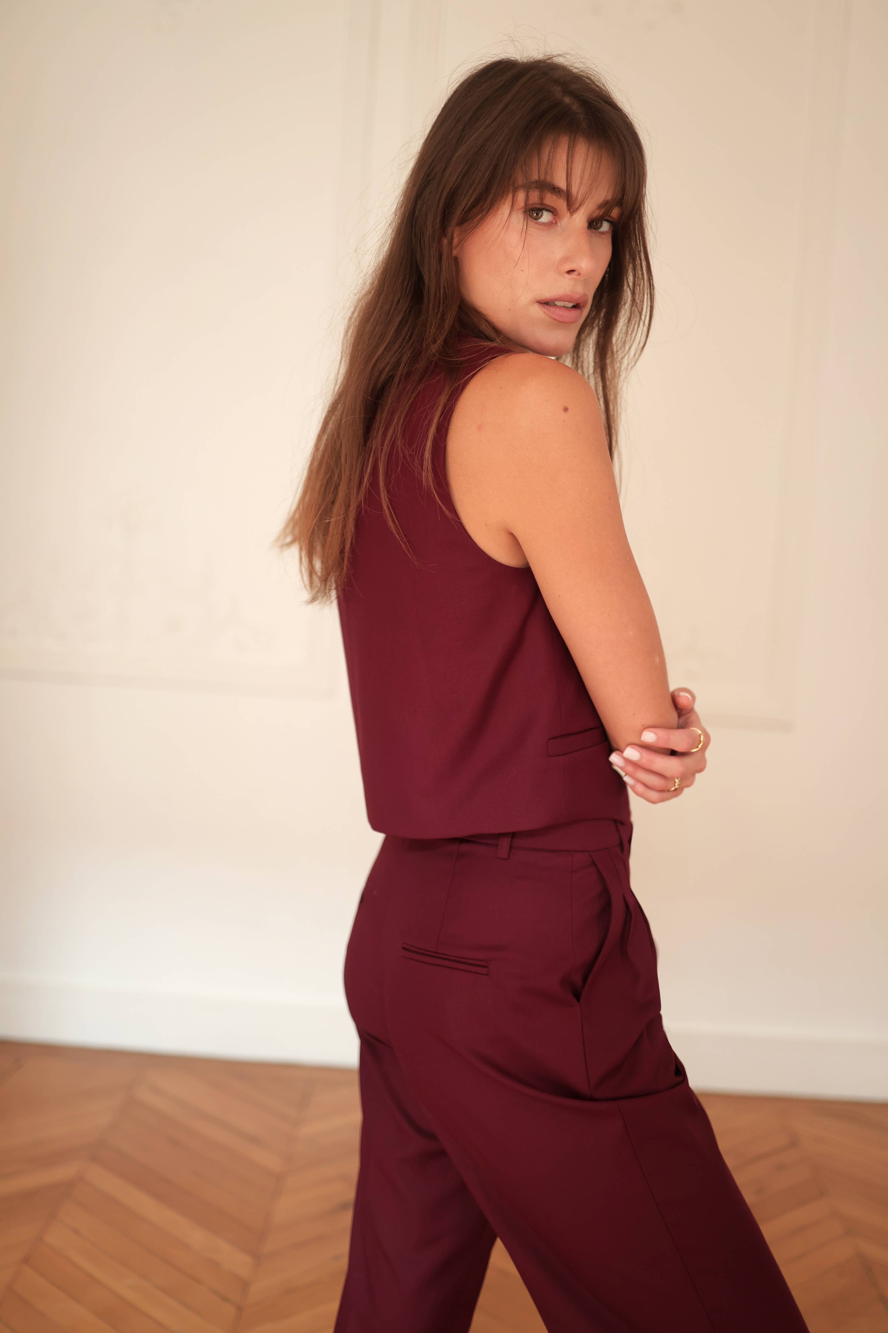 A woman standing against a wall, wearing burgundy sleeveless suit pants with a v-neckline and front pleats.