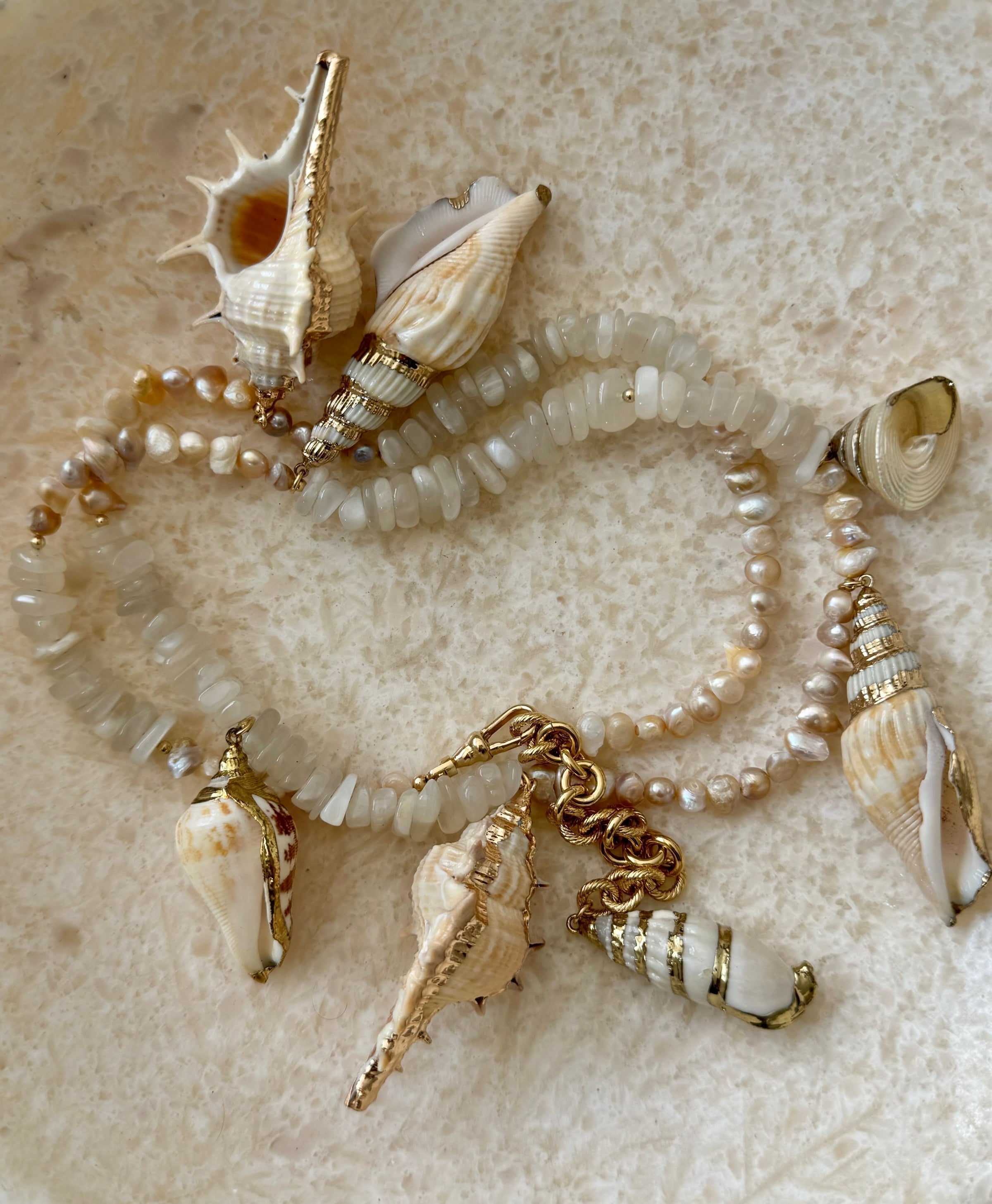 A beaded bracelet featuring white and cream faux pearls, translucent white beads, and multiple natural seashell charms with gold-tone hardware, displayed on a textured beige surface.