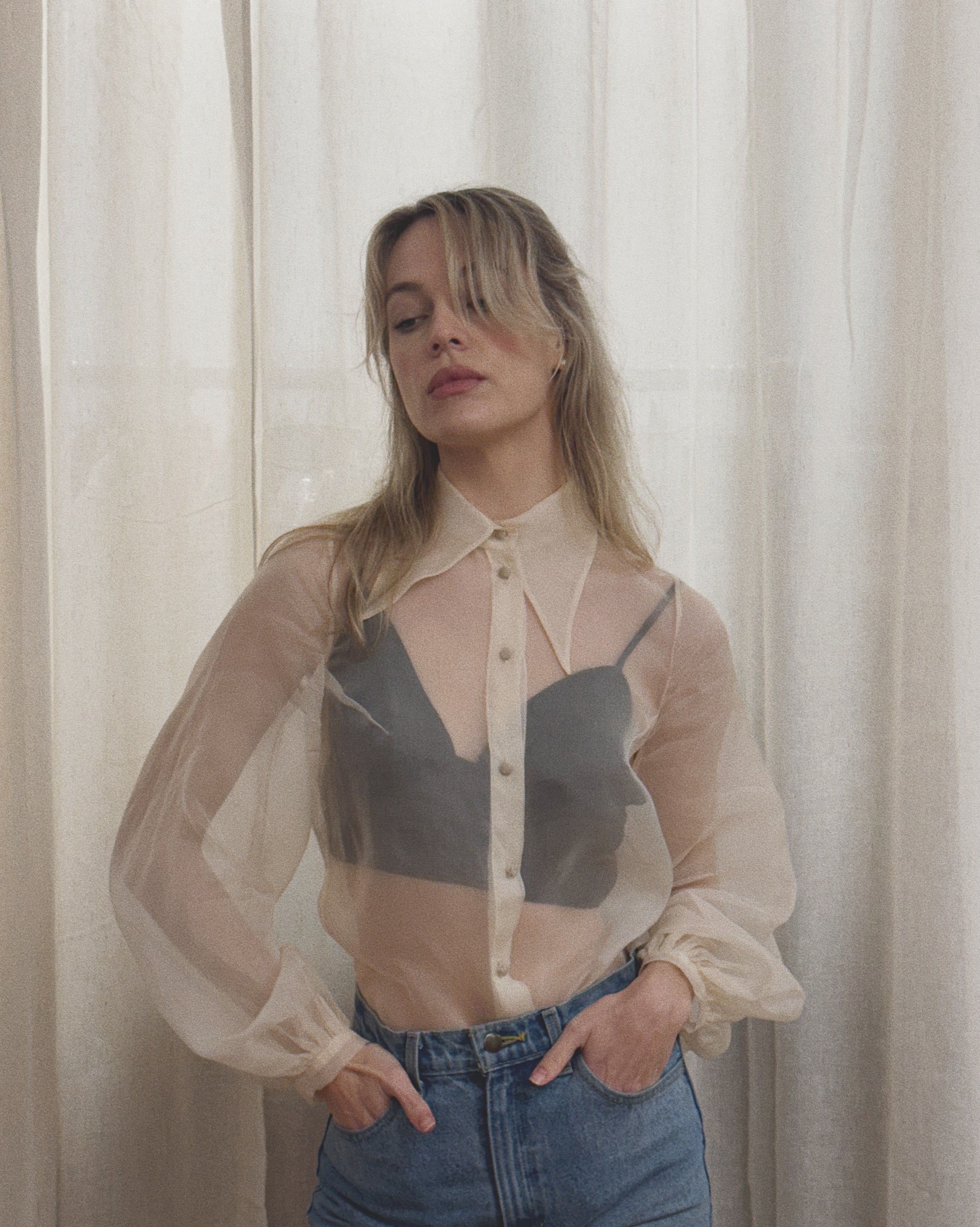Woman wearing a sheer beige blouse over a black top with blue jeans against a white curtain background