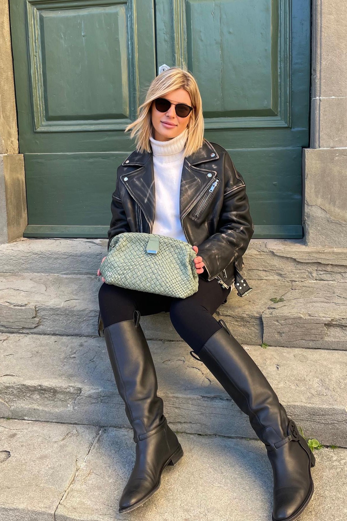 Woman sitting on steps wearing a black leather jacket, white sweater, and black knee-high boots, holding a green handbag.