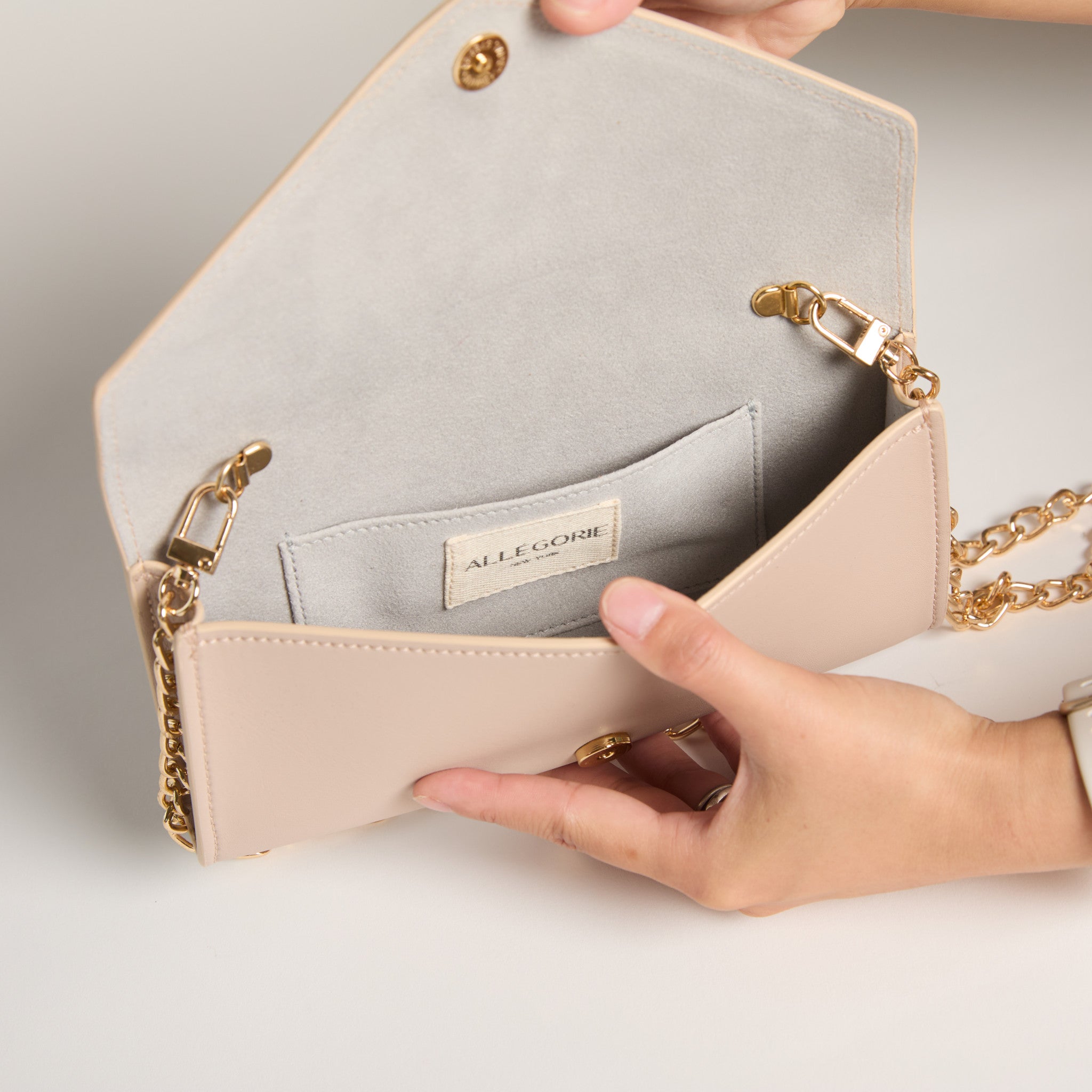 Beige handbag with gold chain detail held by a hand, brand 'ALLEGORIE' visible.