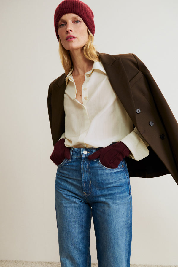 Woman wearing a brown coat, white shirt, blue jeans, and red beanie against a plain background | Exclude